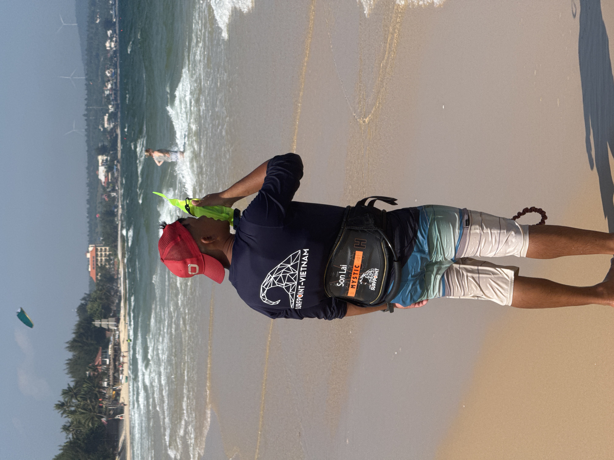 In the coastal city of Mũi Né, Vietnam, a man stands on a sandy beach, facing away from the camera, holding a bright green object to his mouth. He wears a navy blue long-sleeved shirt with a white geometric wave design and the text "SURFPOINT-VIETNAM" on the back, a red baseball cap, and patterned board shorts. A black waist pouch with "SON LAI", "MYSTIC", and "SURFPOINT" logos is secured around his waist. The ocean, with moderate waves, stretches out before him, and in the distance, buildings, palm trees, and wind turbines are visible. A kite is visible in the sky. Another person, a woman wearing a patterned dress, stands in the shallow water.