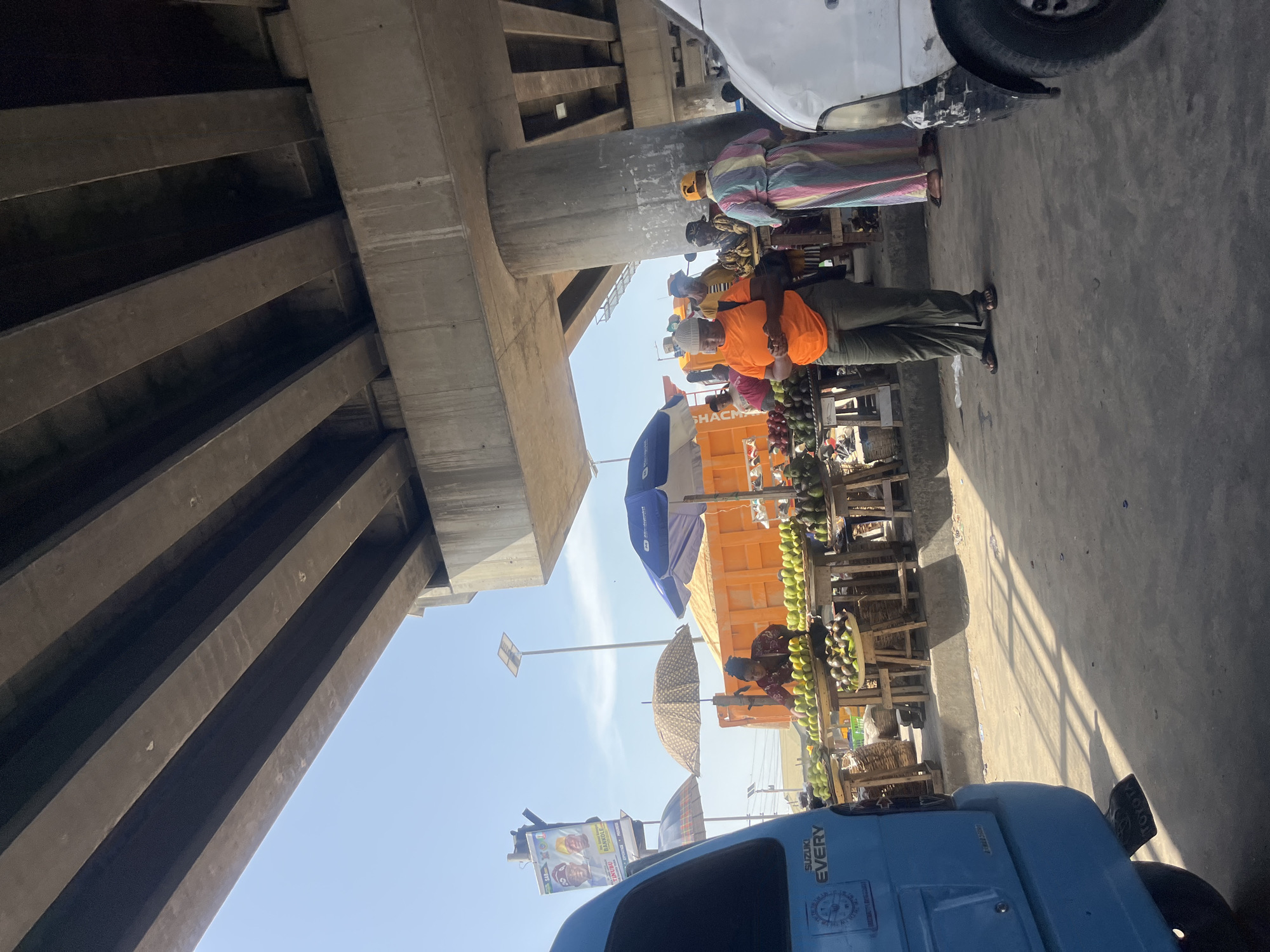 The scene depicts a bustling market environment under an overpass in Ado, Nigeria. The overpass itself dominates the top-left quadrant of the frame, its concrete beams casting shadows. Below, vendors display their wares, mainly fruits such as avocados and mangoes, arranged on wooden stalls. Several people are present, including vendors and customers, with some individuals wearing colorful clothing. A blue Suzuki "Every" mini-truck is parked at the bottom. The light suggests it's daytime. Signboards and umbrellas are visible amongst the market stalls. A white vehicle is parked at the top. The ground appears to be concrete.