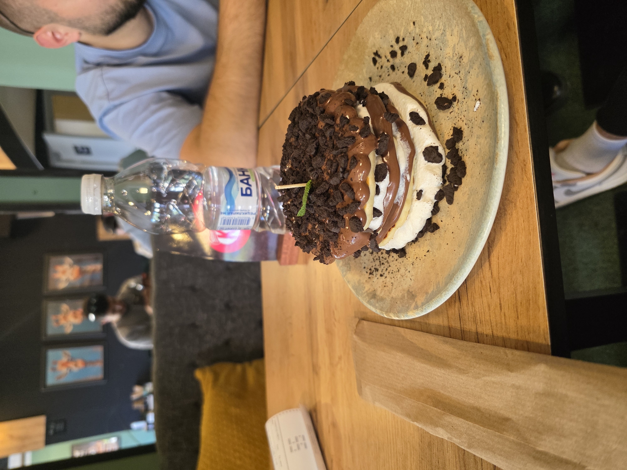 A photo of a dessert on a plate, a bottle of water, and a person at a table in a restaurant, presumably in Veliko Tarnovo, Bulgaria. The dessert appears to be pancakes layered with cream and chocolate crumbs, served on a patterned plate. Next to the dessert is a bottle of water with Cyrillic labeling. The person at the table is wearing a blue shirt. The background includes wall art and a yellow pillow on a grey chair. The table is made of wood, and there is a brown paper napkin and a receipt on the table.
