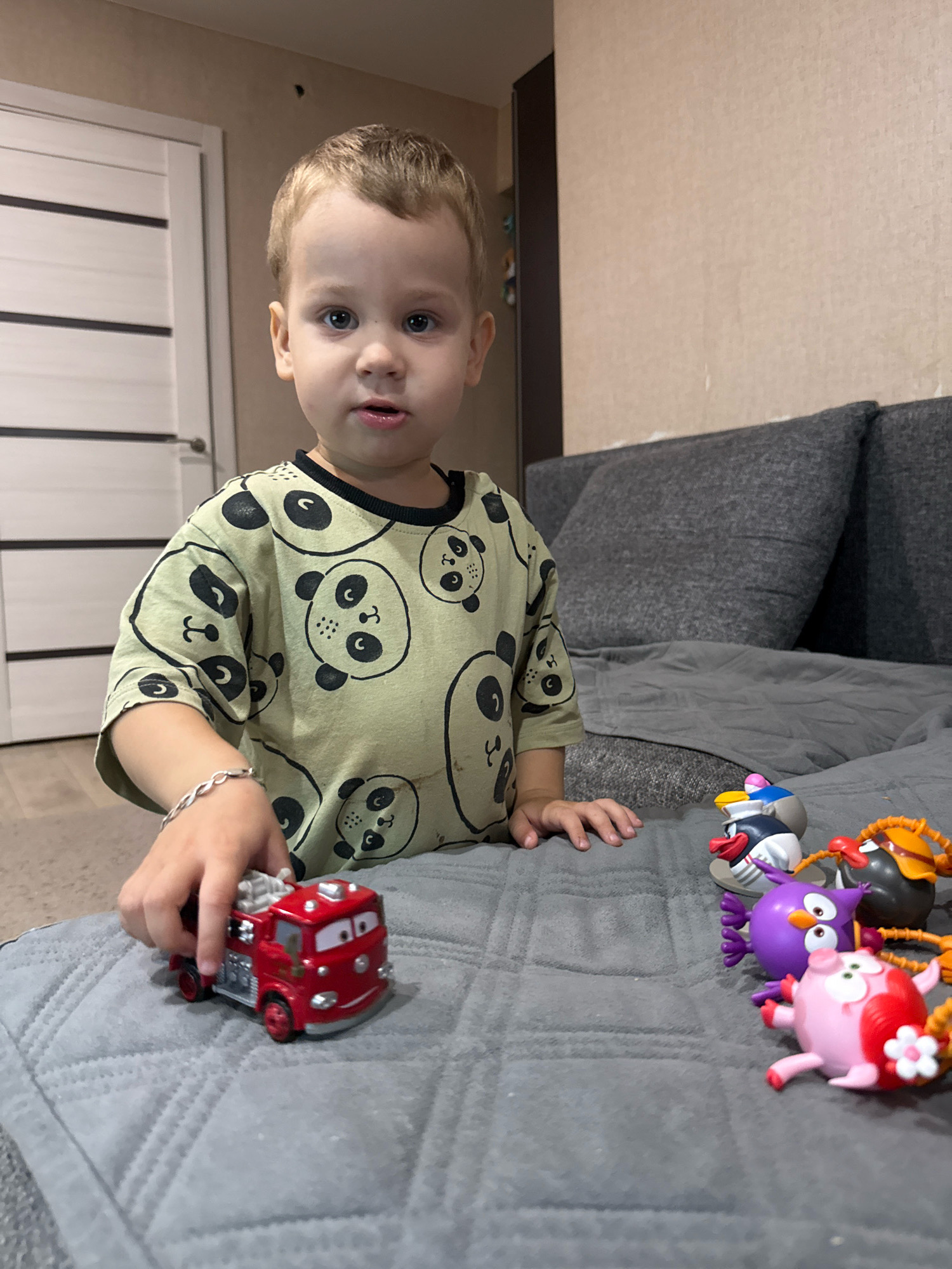A young Caucasian boy with blonde hair and blue eyes, wearing a light green t-shirt with a panda print, sits on a gray quilted surface. He is holding a red toy fire truck with his right hand. To his right, on the same surface, are several small, colorful figurines. The background shows a wall and a dark gray couch with two large cushions. A white door with horizontal lines is visible on the left side of the image. The scene appears to be indoors, possibly in a home environment.