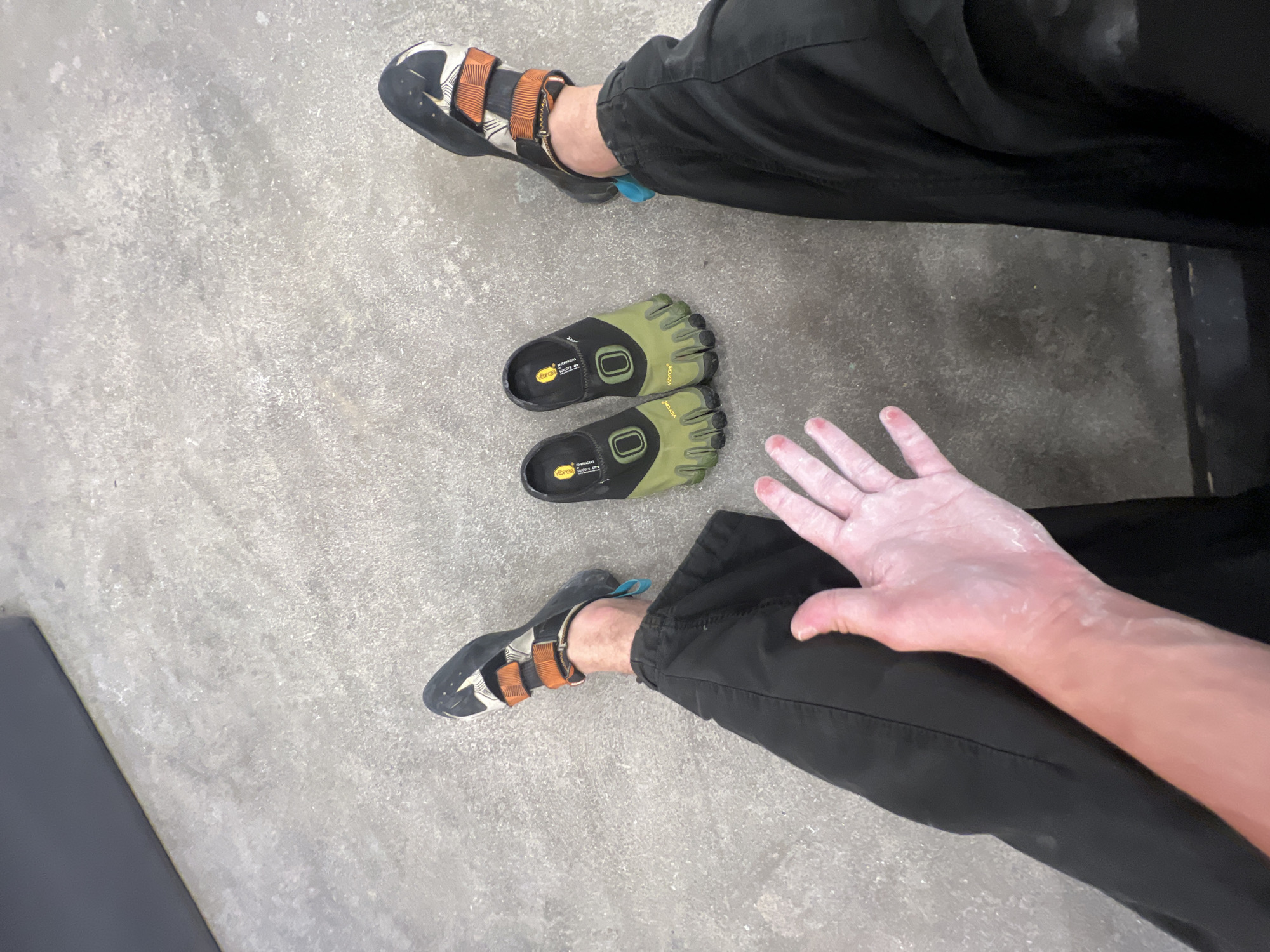 The image shows a person sitting on a concrete floor. They are wearing black pants and climbing shoes. There are two Vibram FiveFingers shoes next to their feet. The person's hands are covered in chalk. The location is likely an indoor climbing gym. There is no information to suggest it is in Sochi, Russia.