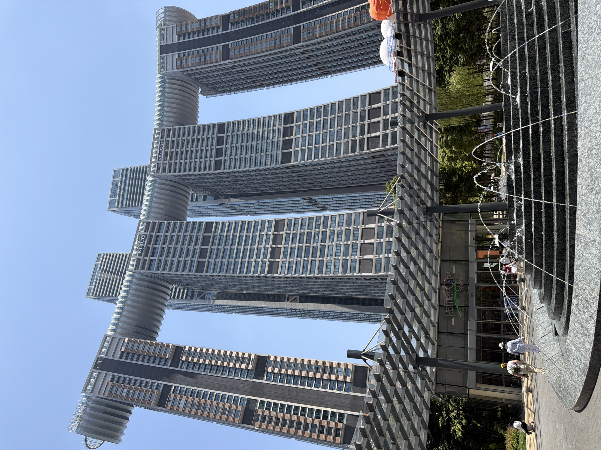 The photograph captures a view of the Raffles City Chongqing complex in the Yuzhong District, China, on a clear, sunny day. The iconic structure consists of four interconnected skyscrapers, with the upper floors of two of the buildings linked by a horizontal, wave-like bridge. The skyscrapers are modern and tall, with visible windows and balconies. In the foreground, a tiered fountain sprays water, with several people walking around the area. To the left of the fountain, a person is kneeling on the ground. To the right of the fountain, a glass-covered walkway with a geometric design extends towards the entrance of a building displaying the "Raffles City" logo. Trees and other vegetation are visible behind the walkway. The sky is bright blue, indicating good weather.