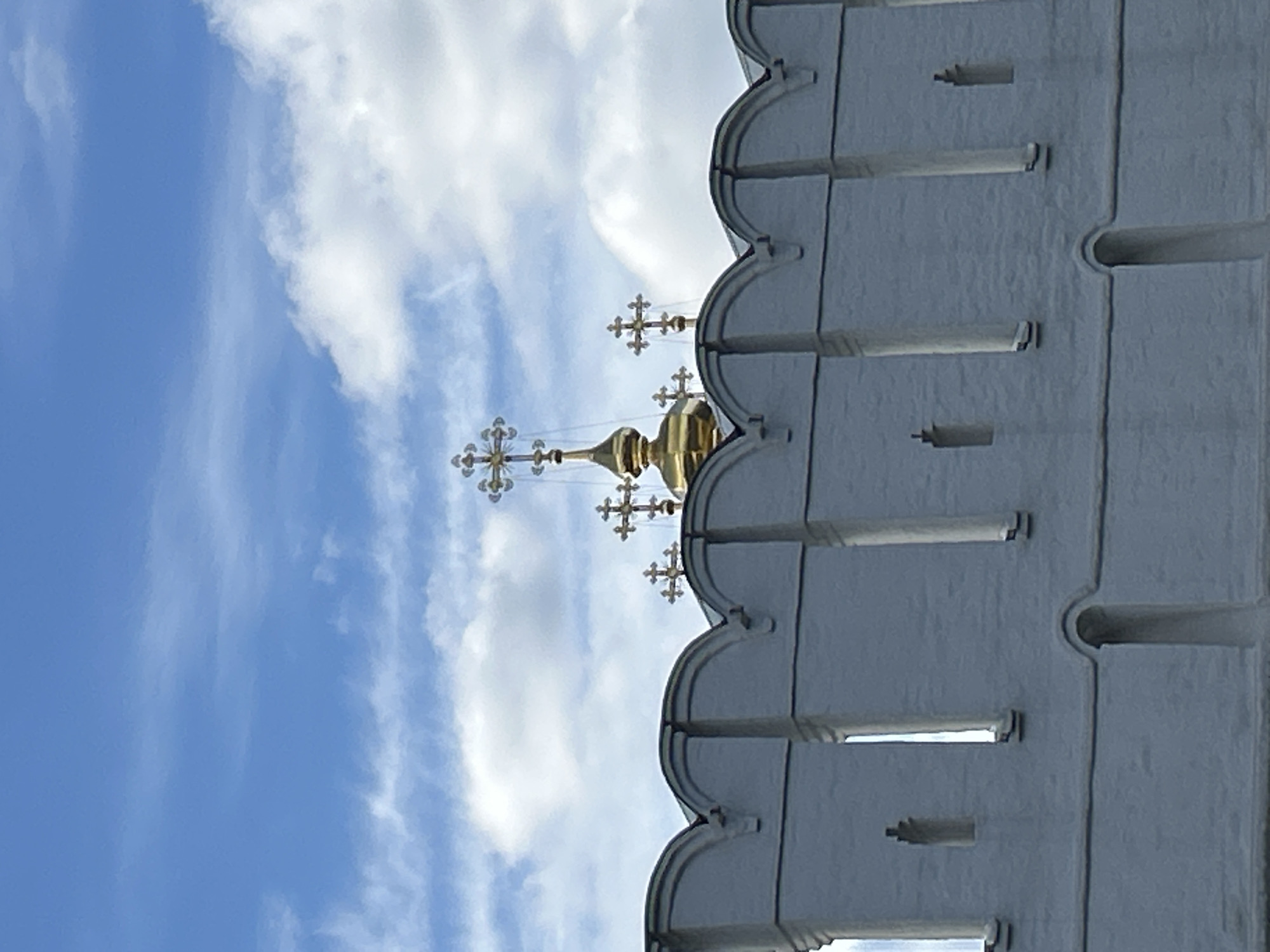 A vertical-oriented, medium shot depicts the golden domes and crosses of a Russian Orthodox church against a backdrop of a blue sky with scattered white clouds. The domes are topped with multiple crosses. Adjacent to the church is a section of a white stone wall, featuring crenellations and vertical support pillars. The scene appears to be in daylight. It is likely the image captures a scene in Moscow, Russia, based on the architectural style of the church and wall.