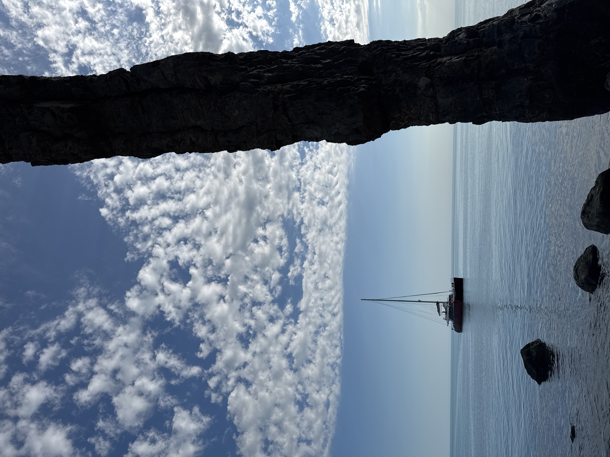 The image captures a serene coastal landscape, viewed through the dark silhouette of a rugged rock formation or natural arch. This massive, craggy rock occupies the upper portion of the frame, creating a strong visual divide. Beyond the arch, the scene opens to a bright blue sky on the left, adorned with numerous scattered, fluffy white clouds (cumulus or stratocumulus) that create a textured pattern.
To the right, a calm, light blue-grey sea stretches towards the horizon under a clearer sky. A distinct red-hulled catamaran sailboat, featuring a tall mast, is visible in the middle distance on the placid water. Its reflection is subtly visible beneath it. In the bottom right corner, a few dark, smaller rocks emerge partially from the water, suggesting a rocky coastline or seabed near the vantage point.
The lighting indicates daytime, likely a bright late morning or early afternoon, with clear weather and gentle sea conditions. The overall mood is tranquil and expansive. The location is Divnomorskoe, Russia, hinting at a Black Sea setting. No people or visible text are present in the image.