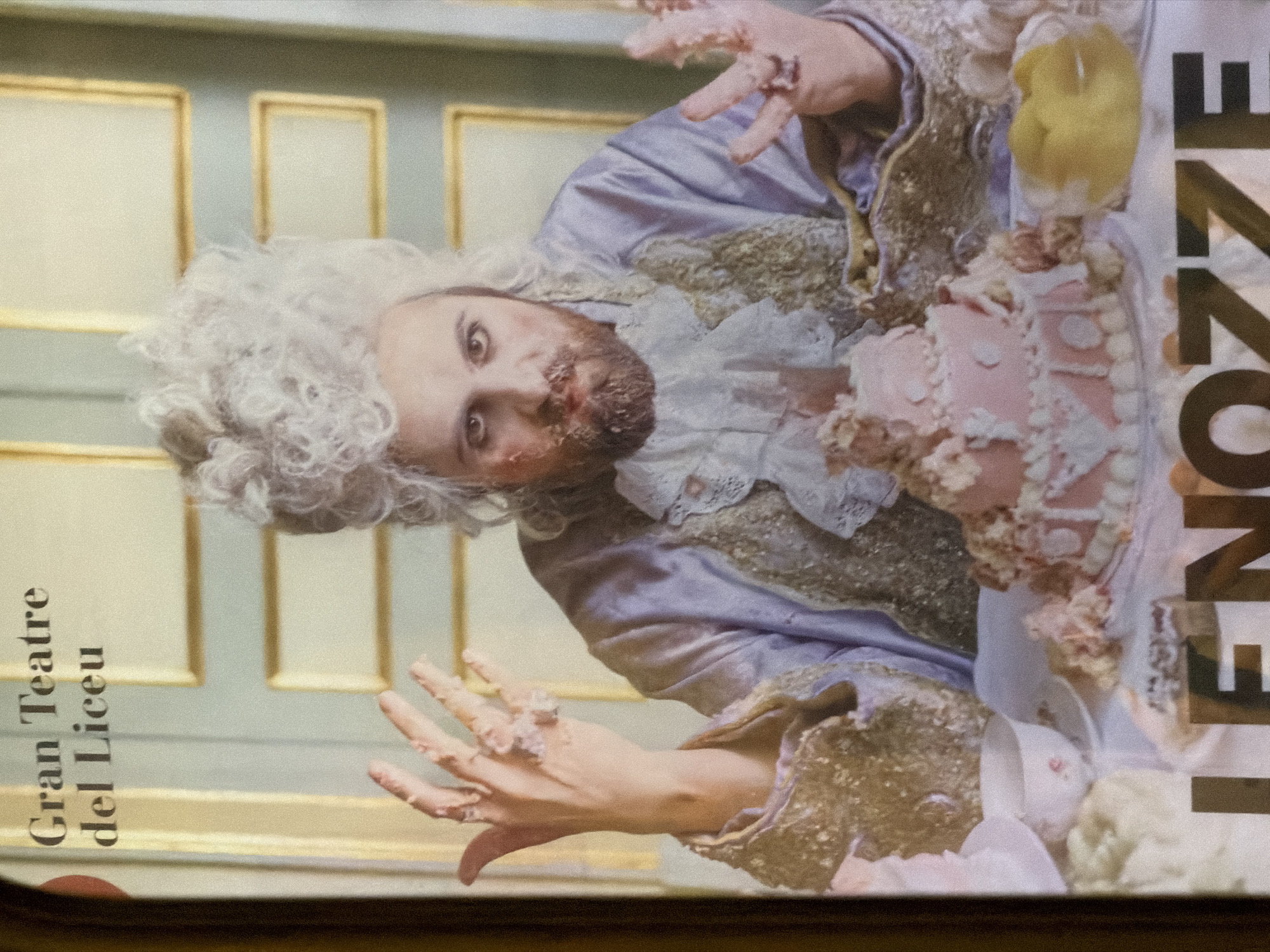 The image is a poster for "Gran Teatre del Liceu" in Barcelona, Spain. It depicts a person in elaborate 18th-century attire, including a white wig and a ruffled collar, with their face and hands smeared with cake frosting. A multi-tiered pink cake sits on a table in front of them, also covered in frosting and crumbs, suggesting a chaotic or perhaps a celebratory event gone awry. The background features a wall with ornate golden rectangular frames, typical of classical architecture. Large black lettering at the bottom of the poster reads "LENOZZZE", likely part of the title of the opera or performance.
