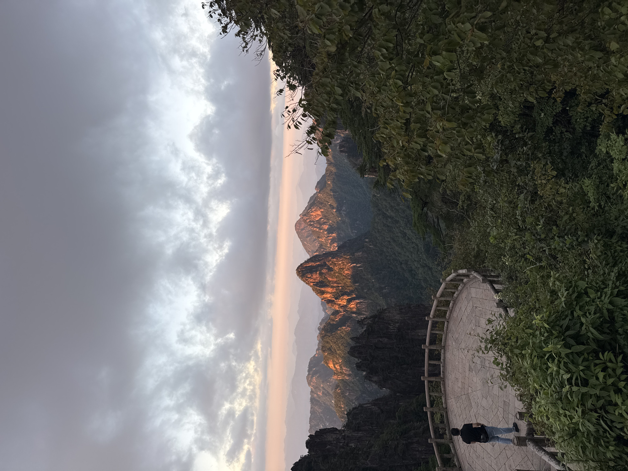 In the city of Huangshan District, China, a lone individual stands on a circular stone observation deck, facing away from the camera and towards a breathtaking mountain vista. The individual, wearing a black t-shirt and blue jeans, appears to be observing the sunset painting the peaks with warm hues of orange and pink. The observation deck, with its stone floor laid in a geometric pattern and a wooden railing, overlooks a vast expanse of lush green mountains and jagged rock formations. The sky above is filled with dramatic, textured clouds, tinged with the soft light of the setting sun. Dense foliage frames the right side of the scene, adding depth and a sense of immersion to the natural landscape.