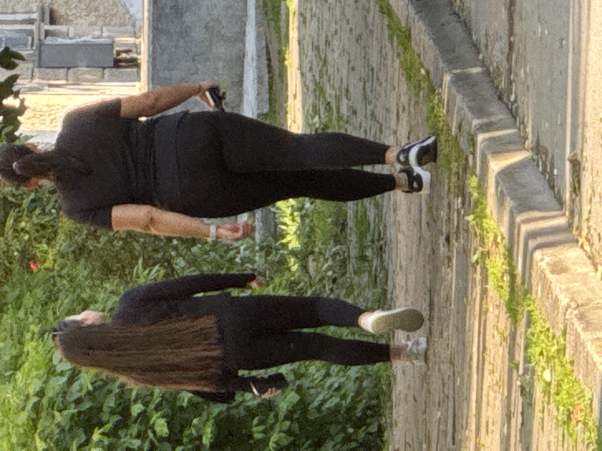 Two women are walking on a paved walkway. One woman is wearing a black top, black leggings, and black and white shoes and is holding a cell phone. The other woman is wearing a black long-sleeved shirt, black leggings, and white sneakers. The walkway is lined with grass and a concrete curb. There is a building in the background with stone accents. Based on the architecture and paving style, this could be in the city of Lovech, Bulgaria, but this cannot be confirmed from the image alone.