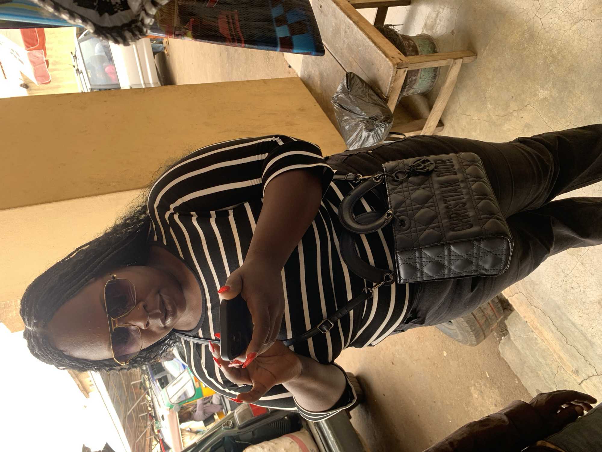 The image captures a person in Bukuru, Nigeria. The individual wears a black and white striped shirt, black pants, and sunglasses, holding a smartphone with red nails visible. A black Christian Dior handbag rests on their lap. The background reveals a partially visible white van, a wooden structure, and a black plastic bag, typical of a roadside environment in Bukuru.