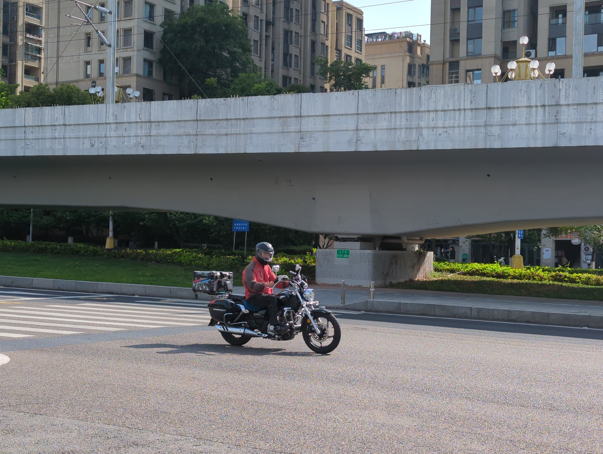 This image depicts an urban street scene in Jiangjin, China, featuring a motorcyclist under a large concrete overpass on a bright, clear day.
The central subject is an individual riding a black cruiser-style motorcycle on an asphalt road. The rider wears a dark helmet with a clear visor, a reddish-orange jacket, black pants, and gloves. The motorcycle features silver exhaust pipes, chrome details, and multiple storage compartments: two black hard-shell boxes on the sides of the rear wheel, and a larger black and silver box mounted on the rear rack. A white pedestrian crosswalk is visible on the road surface to the left of the motorcycle.
The prominent concrete overpass spans the width of the frame in the mid-ground. Above it, in the background, stand modern high-rise apartment buildings with numerous windows and balconies, interspersed with green trees. Decorative streetlights with multiple globe-like luminaires are visible atop the overpass structure on the right side.
Along the road, sections of green grass and manicured bushes are present. Underneath the overpass, on the left, a blue sign with white Chinese characters reads "轨道维护区域 严禁非法施工," which translates to "Track maintenance area, illegal construction strictly prohibited." On a concrete support pillar for the overpass on the right, a white sign with green text displays "JTX D107-Z003." The number "42" is also faintly visible on one of the background buildings.