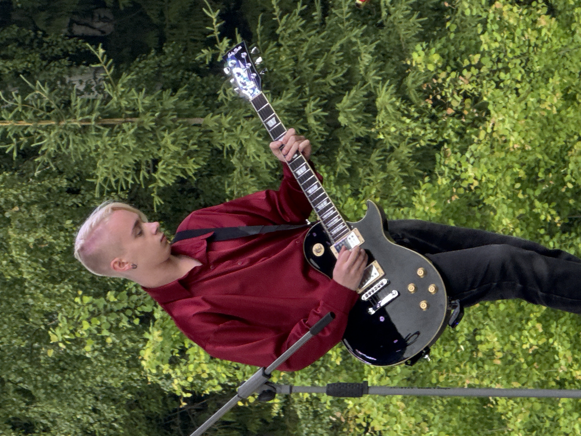 An outdoor daylight photograph depicts a young male individual, estimated to be in his late teens or early twenties, playing an electric guitar. The scene is located in Большая Кивара, Russia. The individual has platinum blonde hair on top with closely shaved sides and is wearing a deep red, glossy, long-sleeved collared shirt and dark trousers. A black guitar strap is visible over his right shoulder. He is actively playing a black electric guitar, identifiable as a Les Paul style, featuring cream-colored pickups, gold control knobs and bridge hardware, and white fretboard binding. The headstock of the guitar is black with a reflective design and text that appears to read "Holy Smoke." His right hand is positioned over the guitar's body near the bridge, and his left hand is on the fretboard. A grey metallic microphone stand with a black adjustment sleeve is visible on the left side of the frame. The background consists of dense green foliage, including various deciduous trees and a prominent slender coniferous tree.