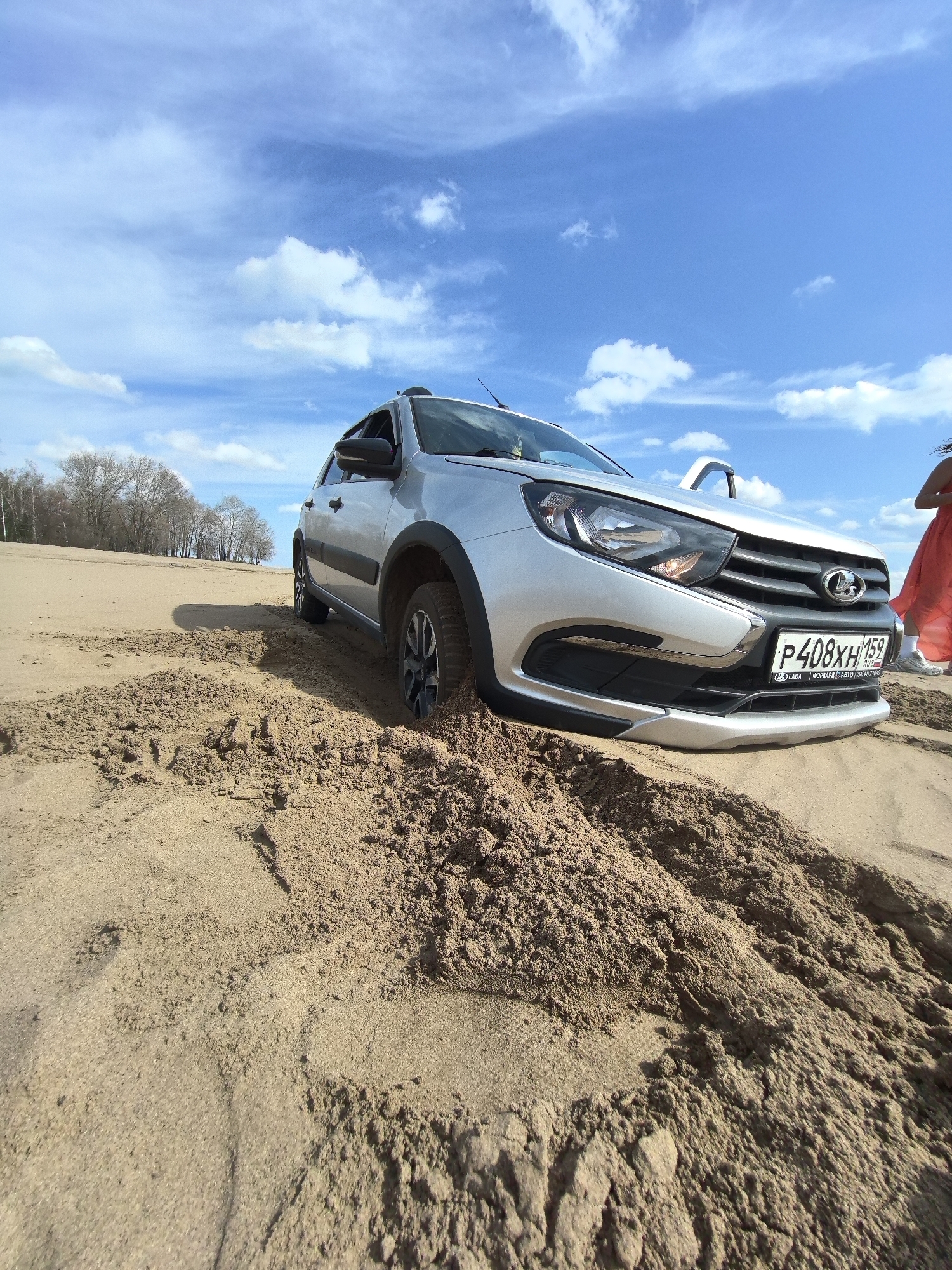 The image depicts a silver Lada Granta Cross car stuck deep in soft, light brown sand under a bright, sunny sky. The vehicle's front right wheel and undercarriage are significantly buried, with mounds of displaced sand surrounding its front section and forming deep ruts from its tires.
The sky is a vibrant blue with scattered white cumulus clouds. In the background, a line of leafless deciduous trees stands on a slightly elevated sandy bank, suggesting a natural, possibly riverside or lakeside, sandy environment. The time of day is clearly daytime, given the bright illumination.
A person wearing a flowing red or orange garment is partially visible on the far right of the frame, standing near the car's front. The front passenger door of the car is open. The Russian license plate, "P408XH 159," is clearly legible on the front of the car. Text on the license plate holder reads "LADA" and "ФОРВАРД АВТО (342) 201 41 40" (FORWARD AUTO).
The scene captures a moment of a car being stranded, likely from an attempt to drive on the challenging sandy terrain. The location is Chaikovsky, Russia, as indicated by the Perm Krai region code '159' on the license plate.