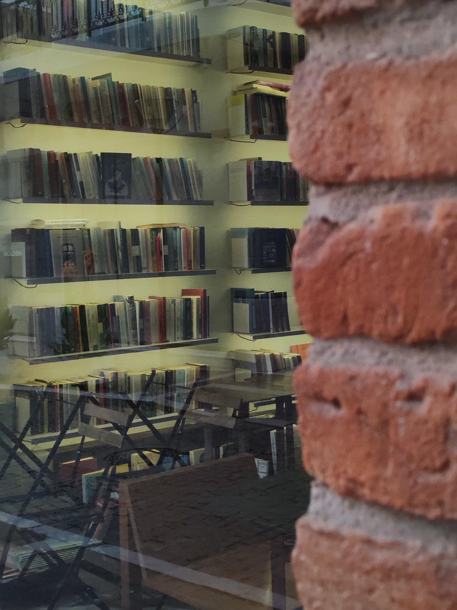 The image shows a view through a window of what appears to be a bookstore or library. Shelves of books are visible on the left side of the frame, arranged in several rows. The books have various colored spines, suggesting a diverse collection. Some of the books appear to be old, with some even showing signs of wear. In the foreground, on the right side of the frame, is a close-up view of a brick wall, indicating that the window is set within a brick building.
Through the window, beyond the shelves of books, there are reflections of tables and chairs, suggesting an indoor seating area. The lighting appears to be artificial, giving the interior a warm glow. The overall impression is one of a cozy and well-stocked place for reading and browsing books. The image does not contain any identifiable people or specific actions taking place. The presence of the brick wall and the nature of the interior strongly suggest an urban setting. While the location of Alexandroupolis, Greece, is provided, there are no specific visual cues within the image that definitively confirm this particular city or country.