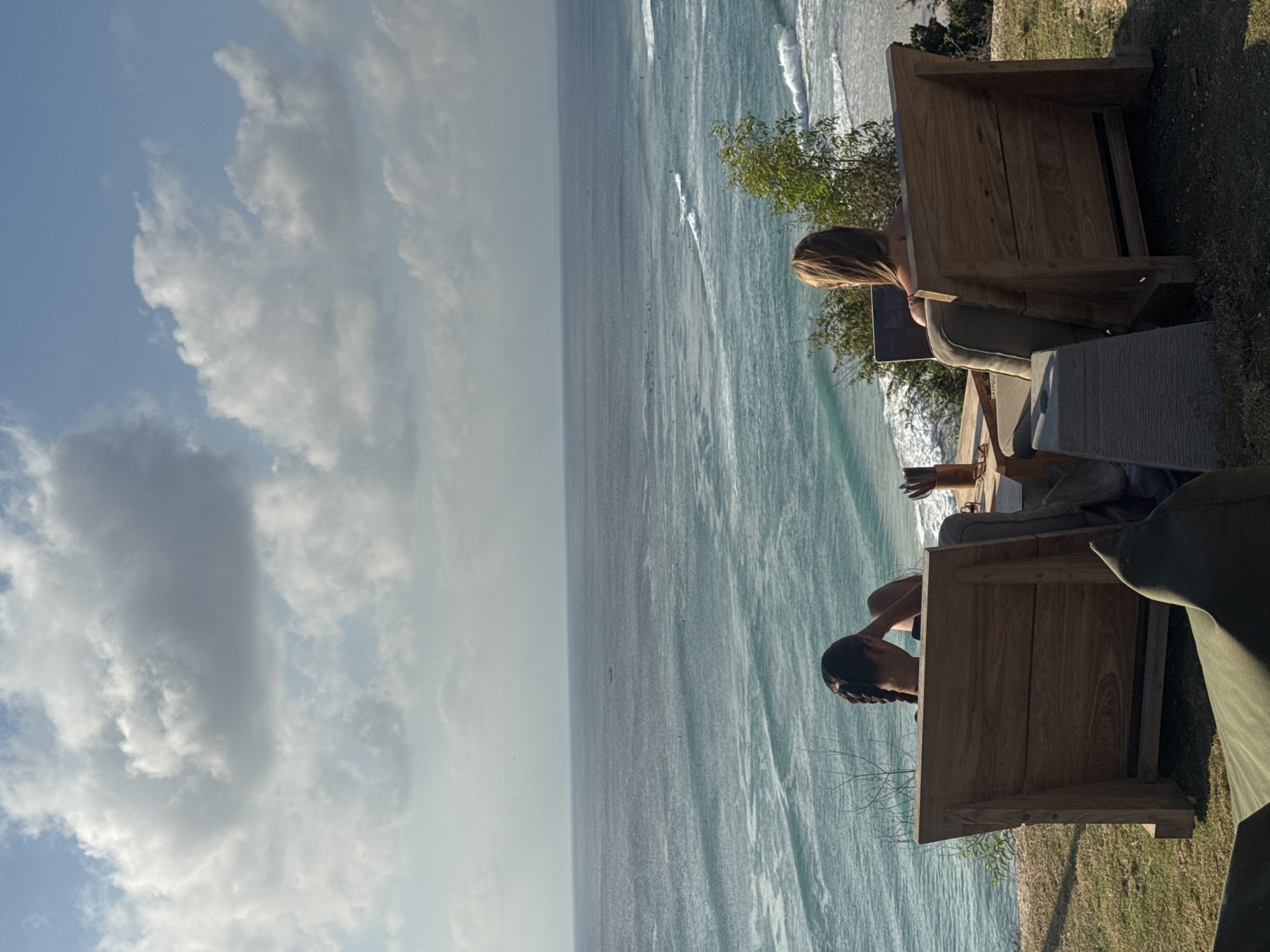 Two women are seated on outdoor lounge chairs facing a vast ocean and sky. The woman on the left, with dark braided hair, is resting her arm on the back of her chair. The woman on the right, with blonde hair, is looking down at a laptop. Both chairs are made of weathered wood and are positioned on a grassy knoll overlooking the water. The ocean is a deep blue with white-capped waves, and surfers are visible in the distance. The sky above is a clear blue with scattered white clouds. The scene is set in Kuta Selatan, Indonesia, a popular coastal area known for its beautiful beaches and surf spots.
