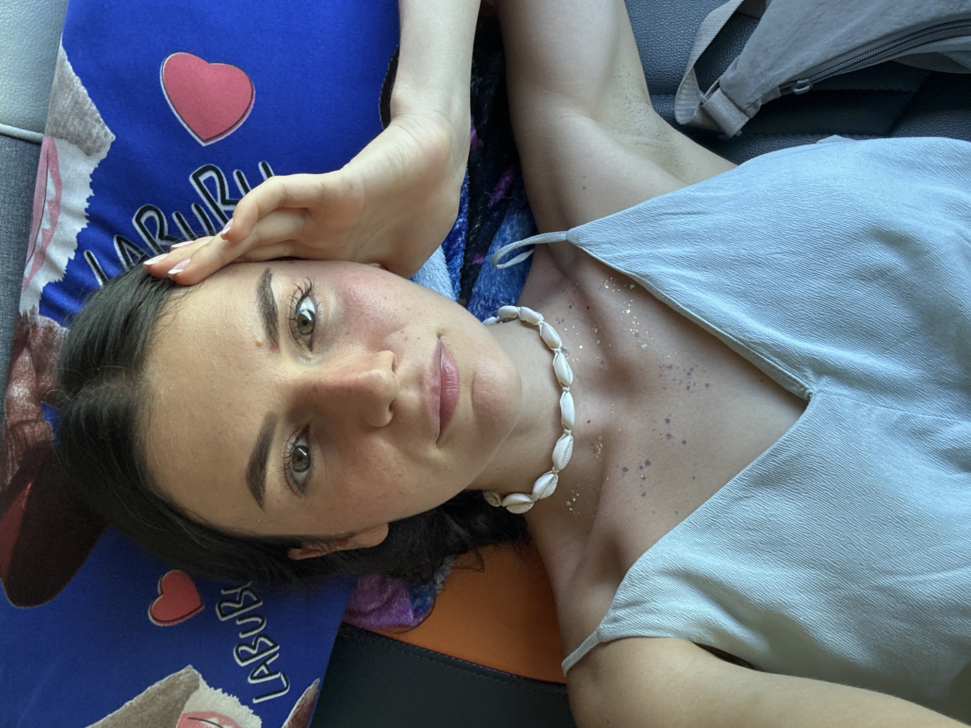 A woman lies on what appears to be a vehicle seat, possibly a car or bus, judging by the visible headrest and seat material. She wears a light blue top and a seashell necklace. Glitter or small decorations are scattered on her chest and shoulder area. Her face is partially visible, with her right hand resting on her forehead. She has dark hair and light eyes. A blue pillow with cartoon images and a heart design is beside her head. A grey bag with a zipper is in the background. It is not possible to confirm the location in the city of Phu Thuy Ward, Vietnam.