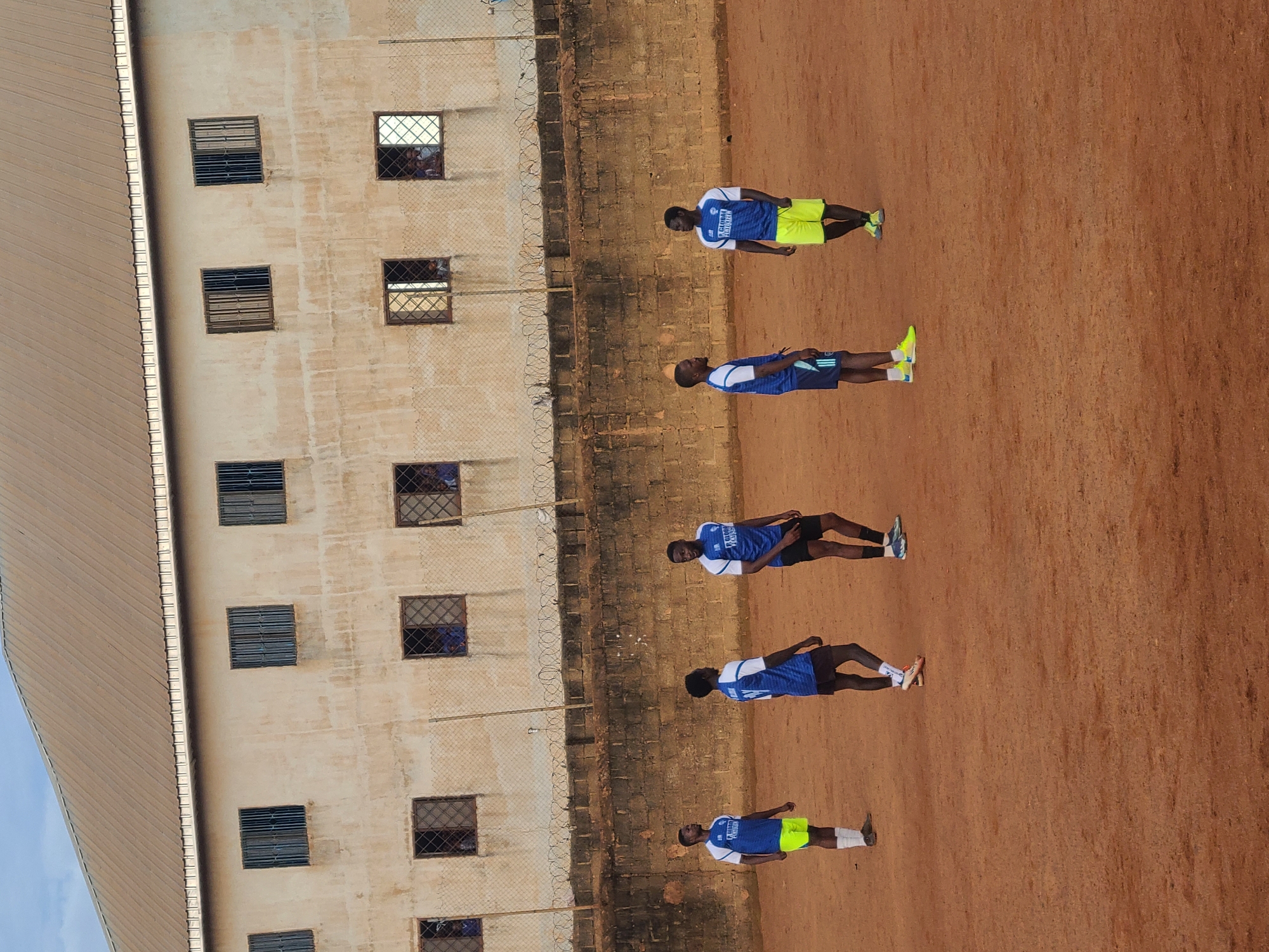 In Barakin Lerawi, Nigeria, five young men in blue and white soccer uniforms are standing on a dusty, reddish-brown soccer field. The field is surrounded by a low brick wall and a chain-link fence topped with barbed wire. Behind the fence is a two-story building with a metal roof, featuring rows of barred windows. Three of the men are wearing blue jerseys with white trim and yellow shorts, one is in blue shorts and a similar jersey, and another is in black shorts and a similar jersey. The men are looking in different directions, some appearing to be in conversation or observing the surroundings. The sky above is a pale blue with some clouds.