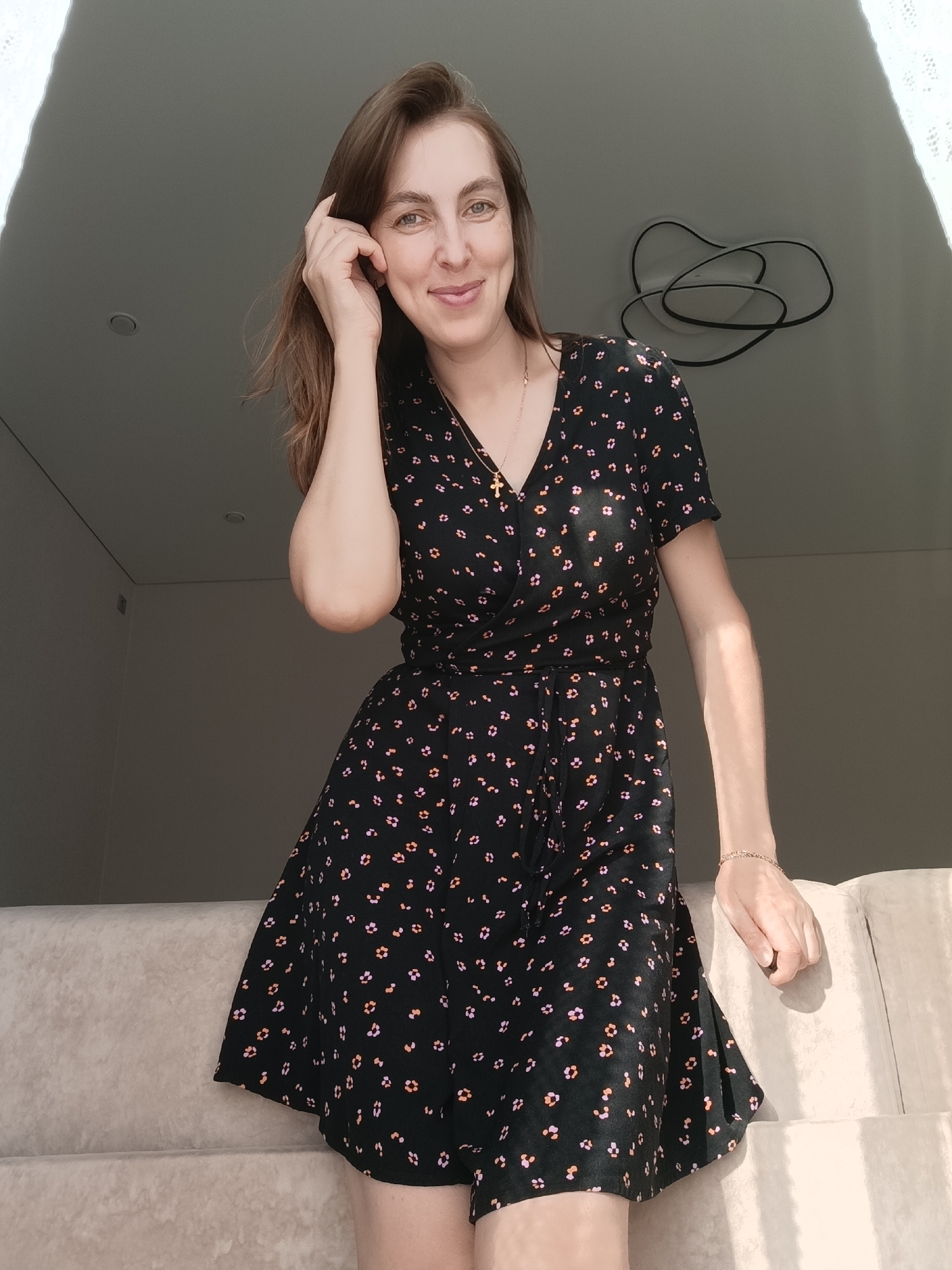 The image depicts a woman in a black floral print dress standing indoors. She is fair-skinned with brown hair, wearing a necklace with a cross and a bracelet. She is smiling and has her hand in her hair. She is leaning against a light-colored upholstered sofa or bench. The background includes a light-colored wall and a ceiling fixture with an abstract design. The lighting appears to be from a nearby window or light source. There is no information available to verify the location of this image to the city of Chaikovsky, Russia.