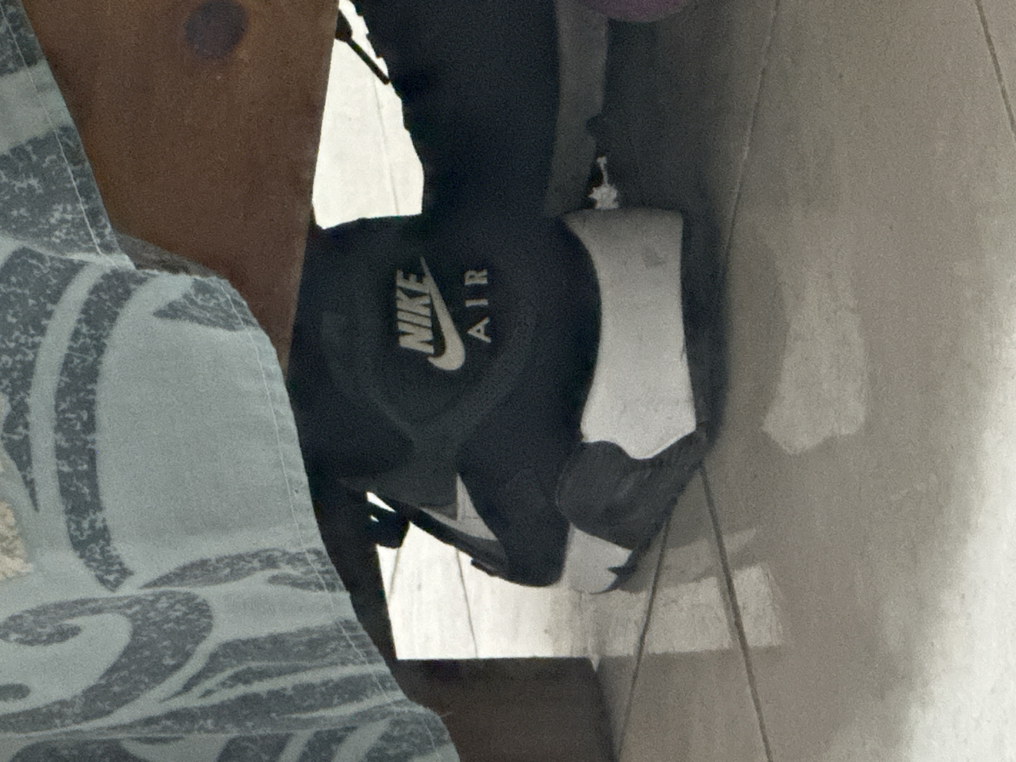 In El Maco, Venezuela, a close-up shot captures a portion of a person's lower leg and foot, partially obscured by a light blue and grey patterned fabric. The individual is wearing a black Nike Air sneaker with a white sole. The sneaker is positioned on a light-colored tiled floor, suggesting an indoor setting. The lighting is subdued, casting shadows on the floor and highlighting the textures of the fabric and the shoe. The scene is static and lacks any obvious action or interaction, focusing solely on the visual details of the clothing and footwear.