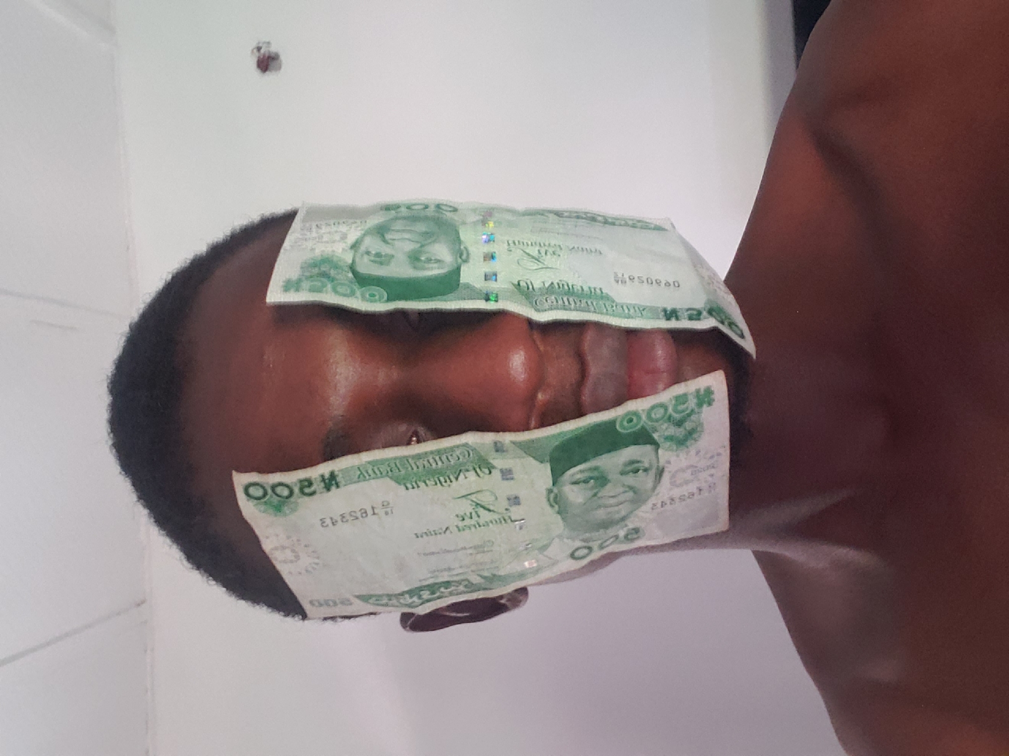 The image displays a person, likely male, with dark skin and short, dark hair, positioned against a plain white wall indoors. The lighting is bright and even, suggesting it is daytime. Two green Nigerian 200 Naira banknotes are placed on the person's face.
One banknote covers the upper part of the face, obscuring the forehead and eyes. This note appears to be placed upside down, as the portrait of Shehu Shagari and the text "TWO HUNDRED NAIRA" are inverted relative to the viewer. The other banknote is positioned across the lower part of the face, covering the mouth and chin, with its portrait of Shehu Shagari and text oriented upright. The act of placing money over the eyes and mouth prevents the viewer from discerning clear facial expressions or emotions, giving the image a potentially symbolic or playful tone.
Visible text on the banknotes includes "CENTRAL BANK OF NIGERIA", "TWO HUNDRED NAIRA", the denomination "₦200", and serial numbers such as "ED58343" and "950580". A small, reddish-brown mark or object is visible on the white wall in the background. The presence of Nigerian Naira banknotes firmly situates the scene in Nigeria, specifically in Adonkolo as per the provided context.