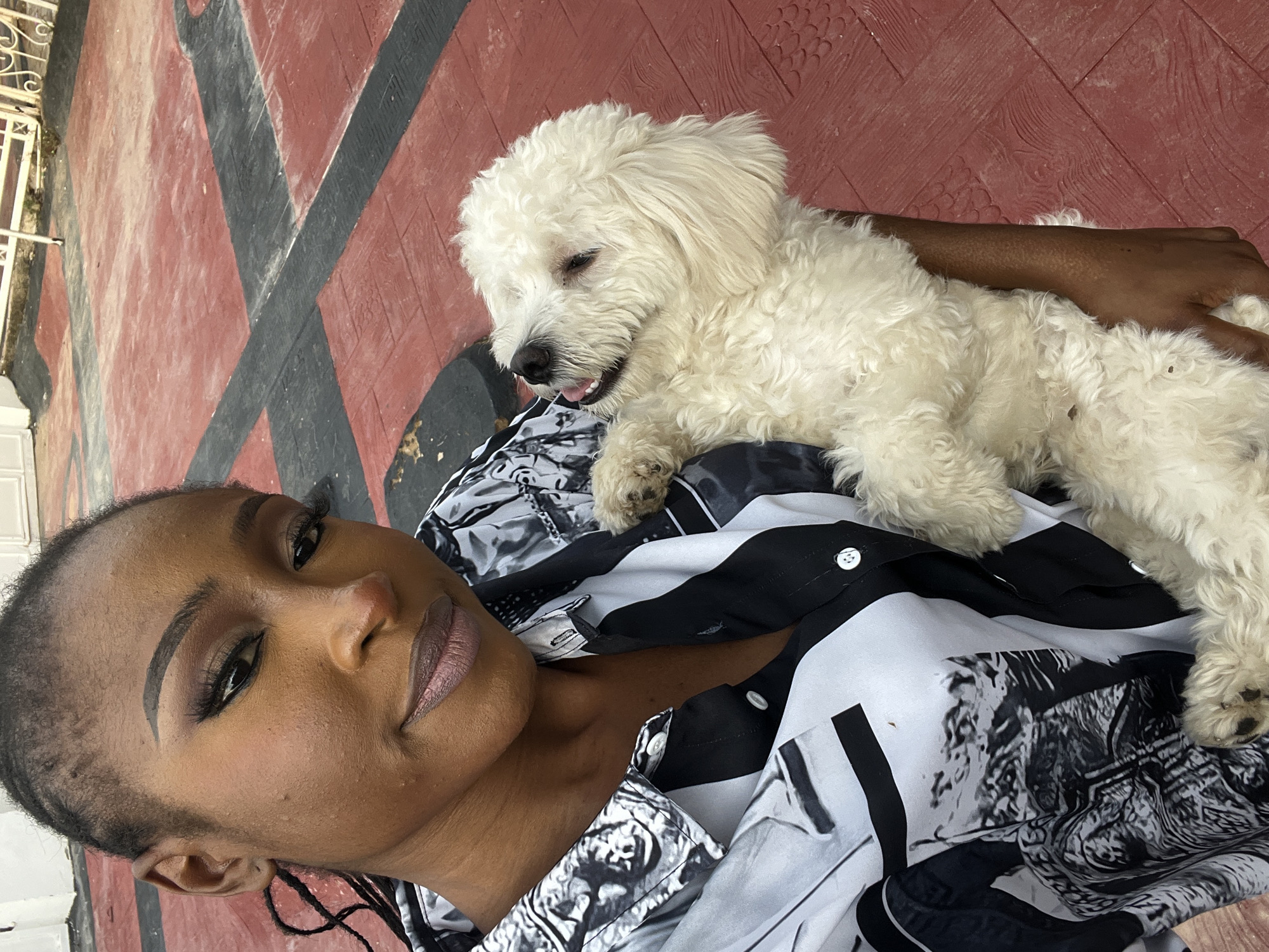 A woman is holding a white dog while standing outside on a red and black tiled floor. The woman is wearing a black and white patterned shirt and has her hair in braids. She is wearing makeup, including eyeshadow and lipstick. The dog appears to be a poodle mix, with fluffy white fur. The background shows a white fence and a bit of greenery. The setting suggests a residential area or a park in Abuja, Nigeria.