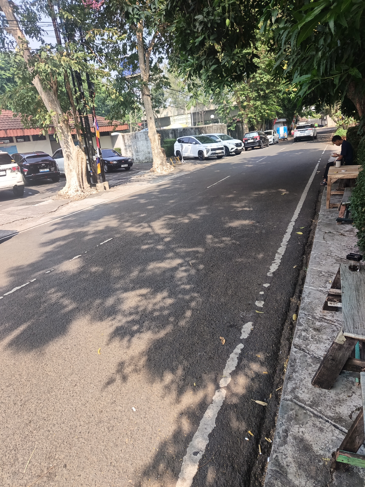 The image shows a street scene in East Jakarta, Indonesia. A road with a white line marking the lane runs through the center of the frame, with trees casting shadows on the asphalt. Several cars are parked on the left side of the road, including white and black SUVs. On the right side of the road, a person is sitting on a wooden bench, looking down at their phone and holding a cup. Next to the bench, there is another wooden structure, possibly a table, with a glass on it. The surroundings appear to be a mix of residential and commercial areas, with buildings and more trees visible in the background. The overall impression is a typical urban street scene during the day.