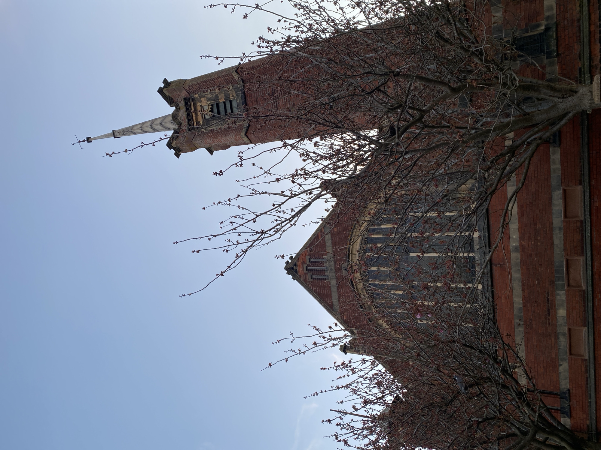 The image features a low-angle view of a red brick building with a tall, pointed spire. Bare tree branches with small buds partially obscure the building's façade. The sky is a light blue, and the photograph is oriented vertically. The building appears to be a church or similar religious structure. It is located in the city of East Suffolk, United Kingdom.