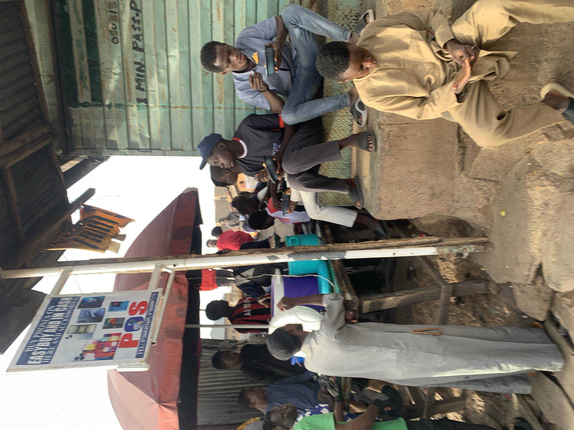 The image depicts a street scene, likely in Jos, Nigeria, based on the presence of a sign advertising "EasyBuy" phones with a local phone number. A group of men is visible, some sitting and eating from plates, while others are standing or walking. The setting includes an array of shops and structures, including an open-air stall with a red awning and various goods on display. The structures around the area include a container, a wooden frame, and a sign with some visible text. The street appears crowded, and there is a mix of clothing styles visible, indicating a typical urban environment.