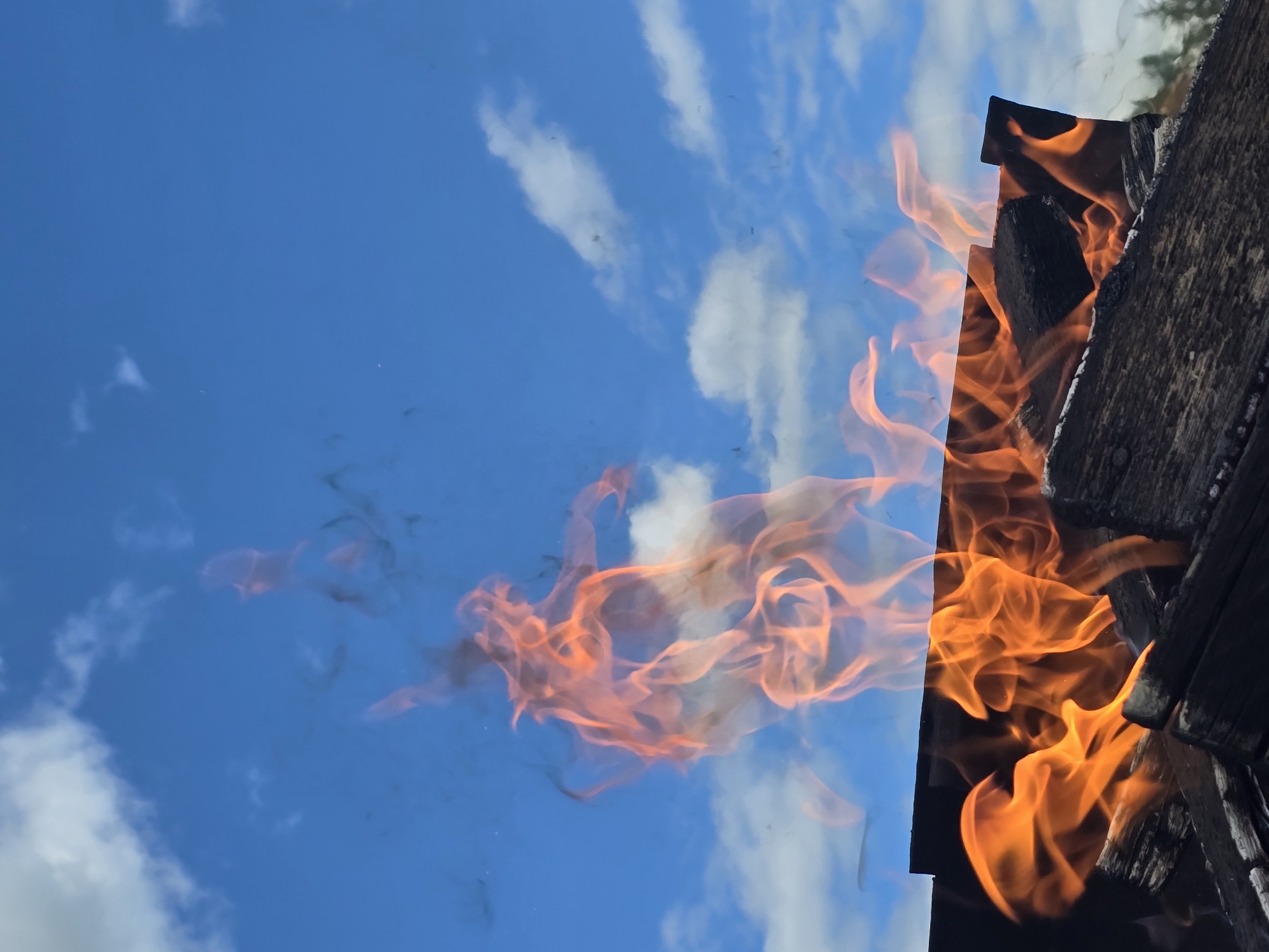 The image depicts a close-up view of a fire burning in what appears to be a fire pit or barbecue grill. The flames are vibrant orange and yellow, licking upwards against a backdrop of a bright blue sky with scattered white clouds. Several pieces of charred wood are visible in the foreground, serving as the fuel for the fire. Wisps of smoke are also present, trailing upwards into the sky. The scene is set outdoors, with a hint of trees in the far distance, suggesting a natural or semi-natural environment. There are no people or animals visible in the frame. The overall impression is one of a bright, possibly warm, day with an active fire. The location is identified as Ермолаево, Russia.