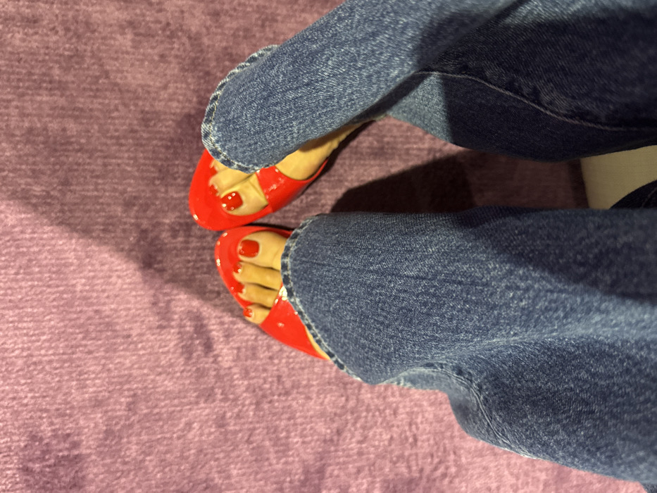 An overhead image displays the lower legs and feet of an individual. The person is wearing blue denim jeans, which are slightly folded or gathered around the ankles. Both feet are clad in bright red, glossy, open-toed flat sandals featuring wide toe straps. The toenails are painted with a matching red polish. The feet are resting on a textured surface of uniform purple color, consistent with carpet or upholstery. A distinct shadow extends from the right side of the frame across the purple surface. The precise location within Dubai, United Arab Emirates, cannot be determined from the provided visual information.