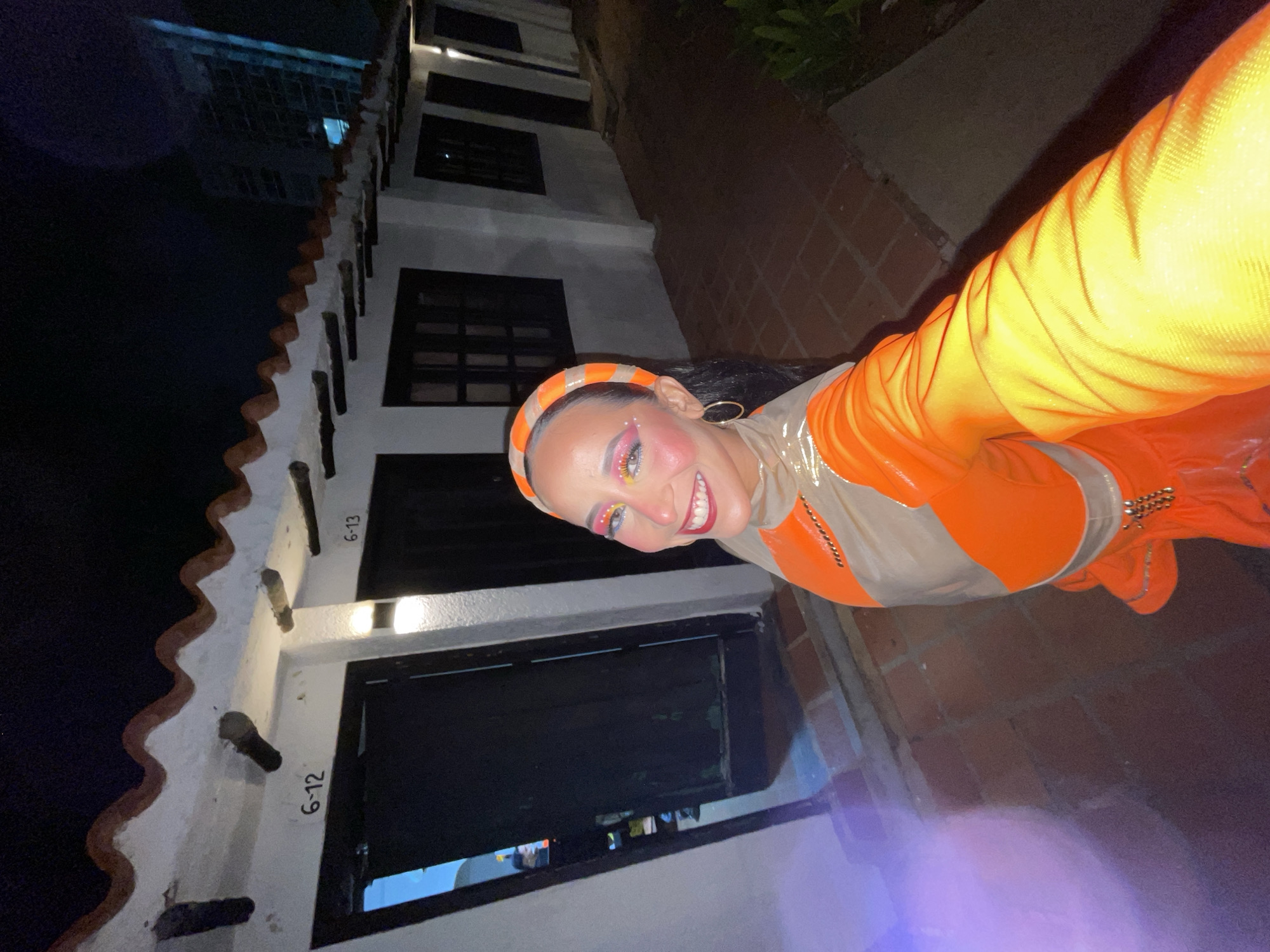 In Porlamar, Venezuela, a woman poses for a selfie outdoors at night. She wears a colorful outfit with orange and tan elements, complete with elaborate eye makeup and an orange headband. The background features a row of white buildings with red-tiled roofs and dark doorways, labeled with room numbers like "6-12" and "6-13". A modern skyscraper is visible in the distance, illuminated against the night sky. The ground is paved with reddish-brown tiles.