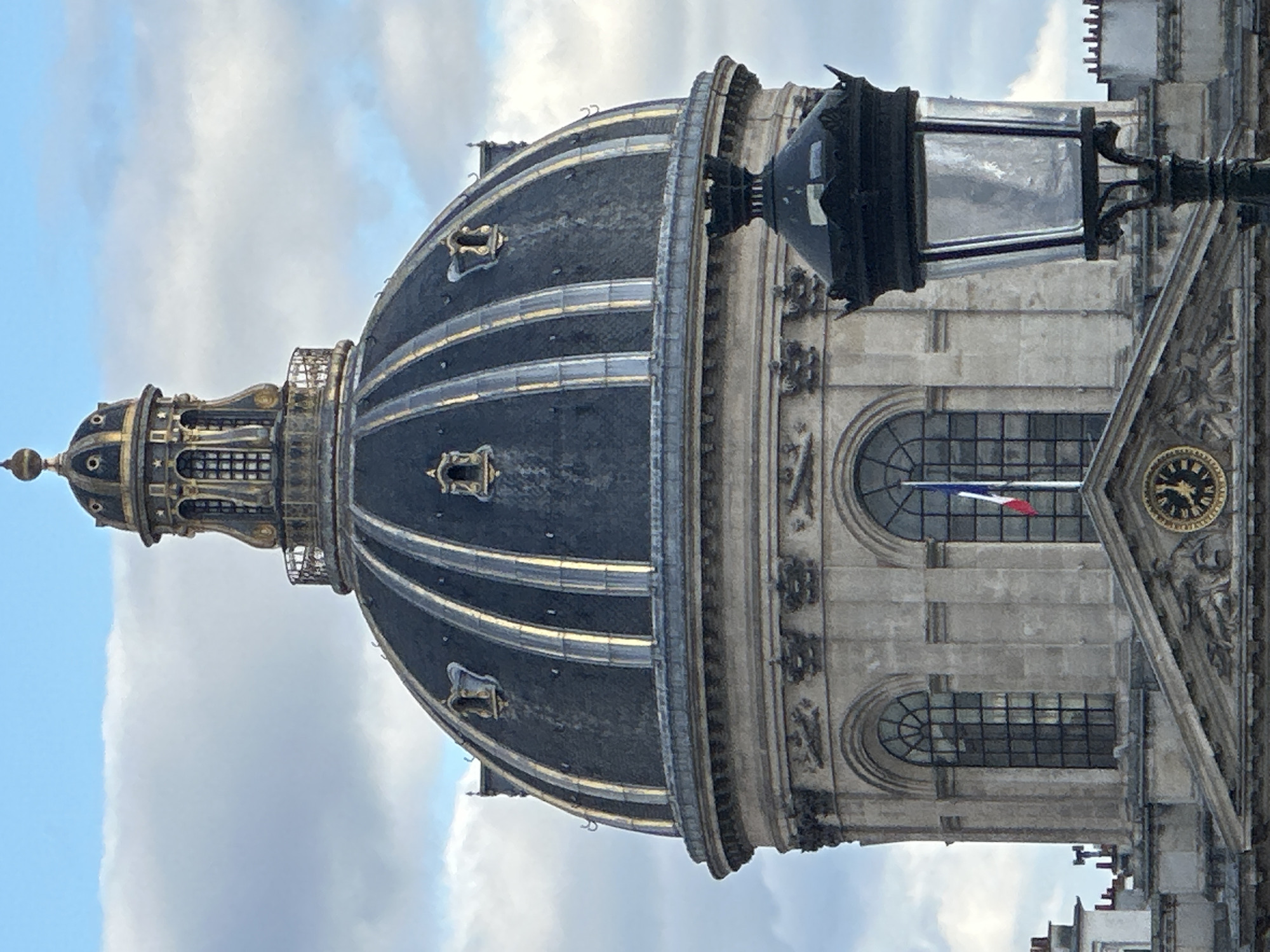The image depicts the dome of the Panthéon in Paris, France. The dome is dark grey with gold accents and is topped with a gilded lantern. A French flag hangs from a window in the building below the dome. A street lamp is visible in the foreground, partially obscuring the view. The sky is cloudy.