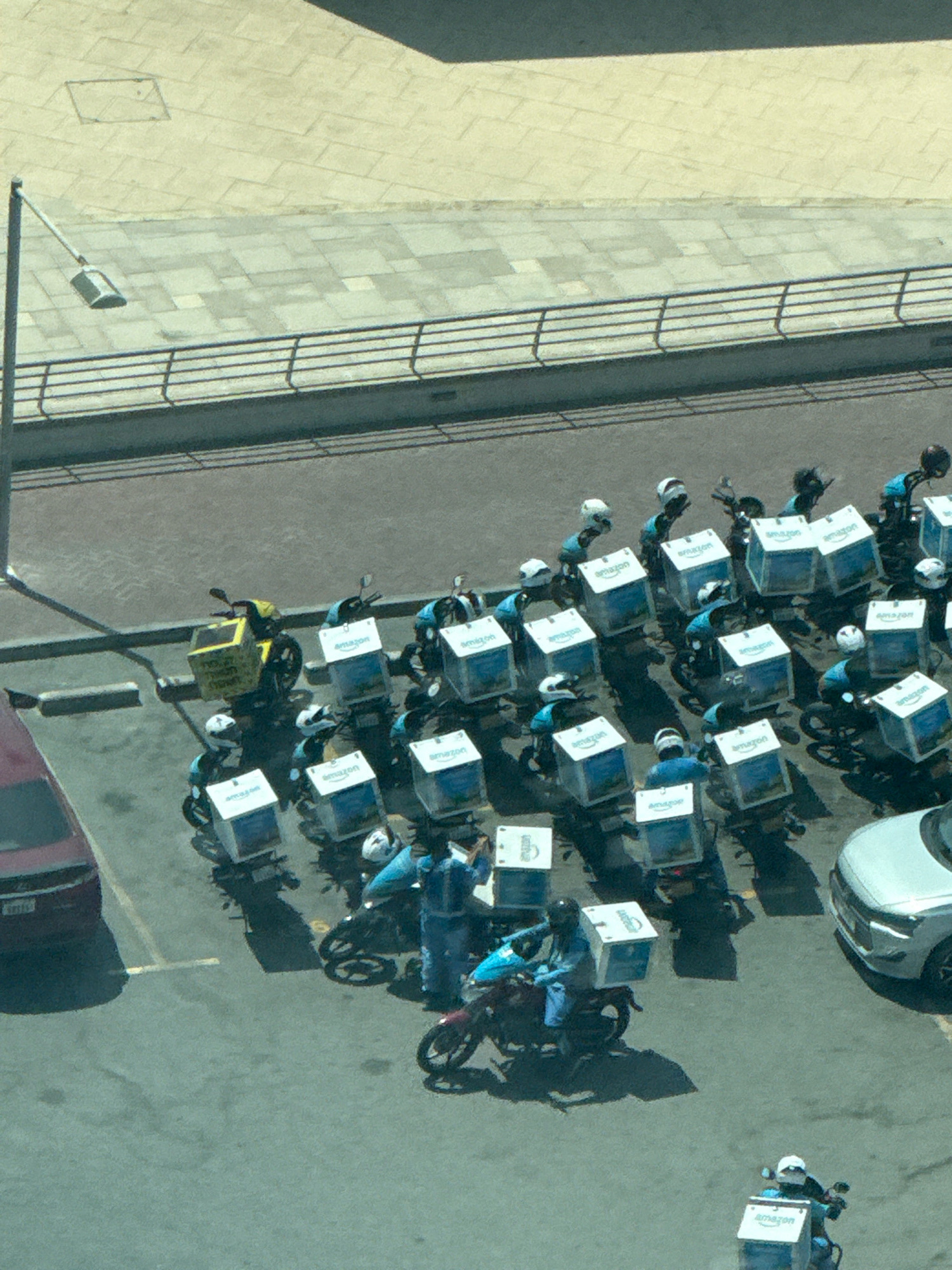 An aerial view captures a parking lot in Dubai, United Arab Emirates, filled with numerous Amazon delivery motorbikes. The scene is illuminated by bright daylight, casting distinct shadows from the vehicles and a nearby lamppost.
The motorbikes, predominantly black with blue and white Amazon branding on their storage containers, are parked in rows. Several riders, dressed in blue uniforms and helmets, are either on their bikes or standing near them, suggesting a gathering or a staging area. One rider is actively engaged with a person in a blue uniform, possibly exchanging packages or receiving instructions. Another motorbike, distinct with a yellow delivery box, is parked separately on the left side of the frame.
A dark red car is parked to the far left of the image, while a silver SUV is visible on the right. The background consists of a paved area with distinct tiling patterns and a metal railing, separating it from a darker paved surface. The overall impression is that of an organized delivery operation in progress.