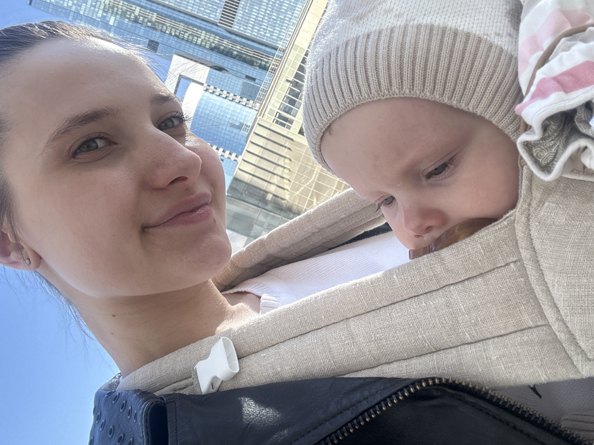 The image captures an adult female holding an infant in a beige fabric baby carrier, set outdoors in a modern urban environment identified as Khimki, Russia. The female, occupying the upper portion of the frame, presents with fair skin, light-colored eyes, and a slight smile while looking directly towards the camera. She wears a black leather jacket with a visible zipper and metal grommets on a shoulder strap, over a white garment, and the baby carrier. A single stud earring is visible in her right earlobe. The infant, positioned in the lower frame, is partially visible in profile, looking towards the left. The infant wears a light beige knitted bonnet with a striped pattern and has a brown pacifier partially inserted in its mouth. A folded textile with pink and white stripes, along with a brown patterned fabric, is visible below the infant's chin within the carrier. The background consists of multiple modern high-rise buildings with reflective glass facades, under a clear blue sky.