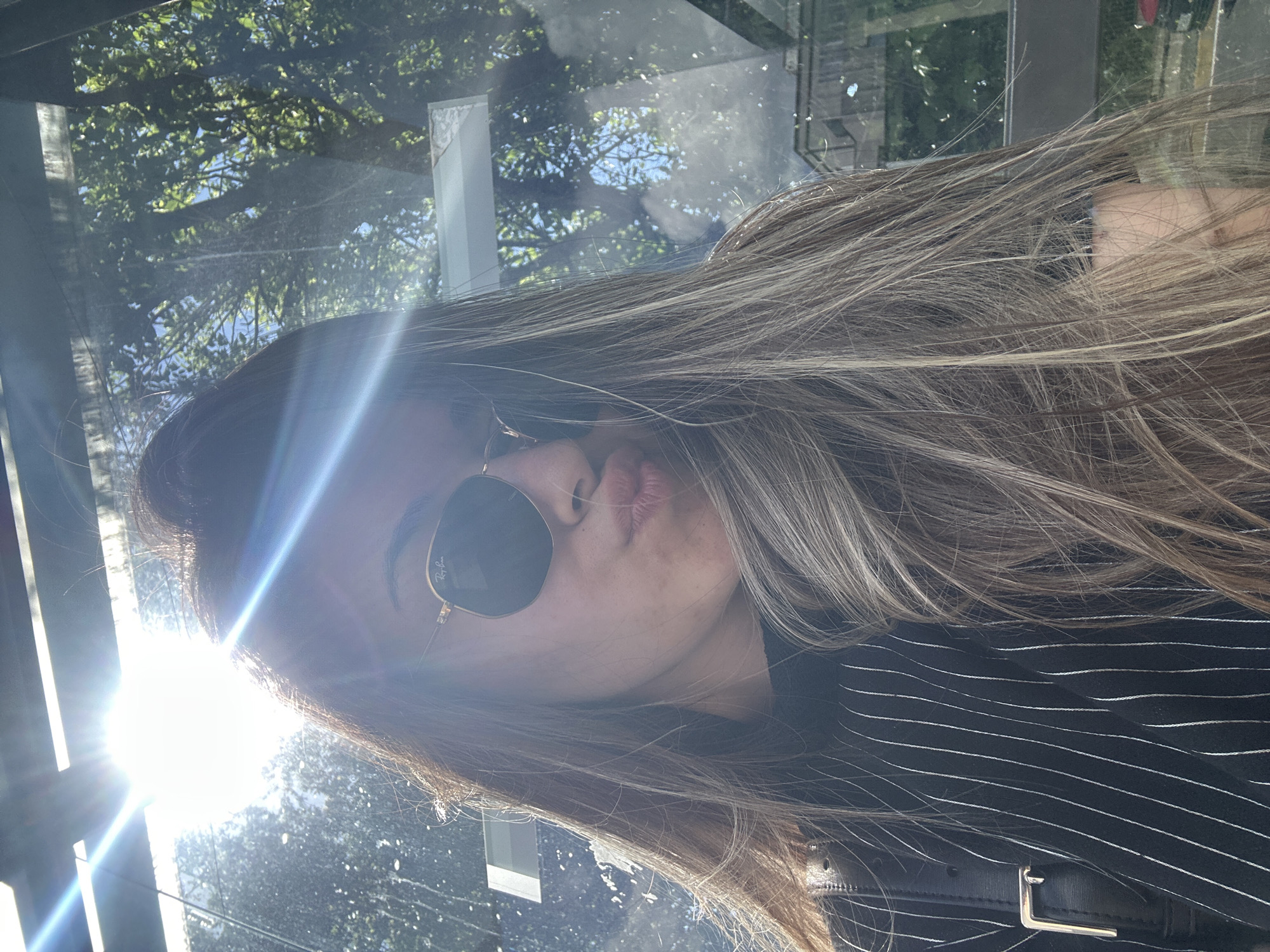 A woman with long, highlighted hair is wearing round sunglasses and a black pinstripe top with a leather bag. She is puckering her lips as if giving a kiss. The sunlight is bright and directly in front of her, obscuring the background. The reflection in the glass suggests trees and buildings, potentially indicating an urban environment. Given the request for a location, and the fact that the user is asking for a location in Sofia, Bulgaria, it is possible that the picture was taken in the bus stop, next to the building, which can be seen in the reflection of the window.