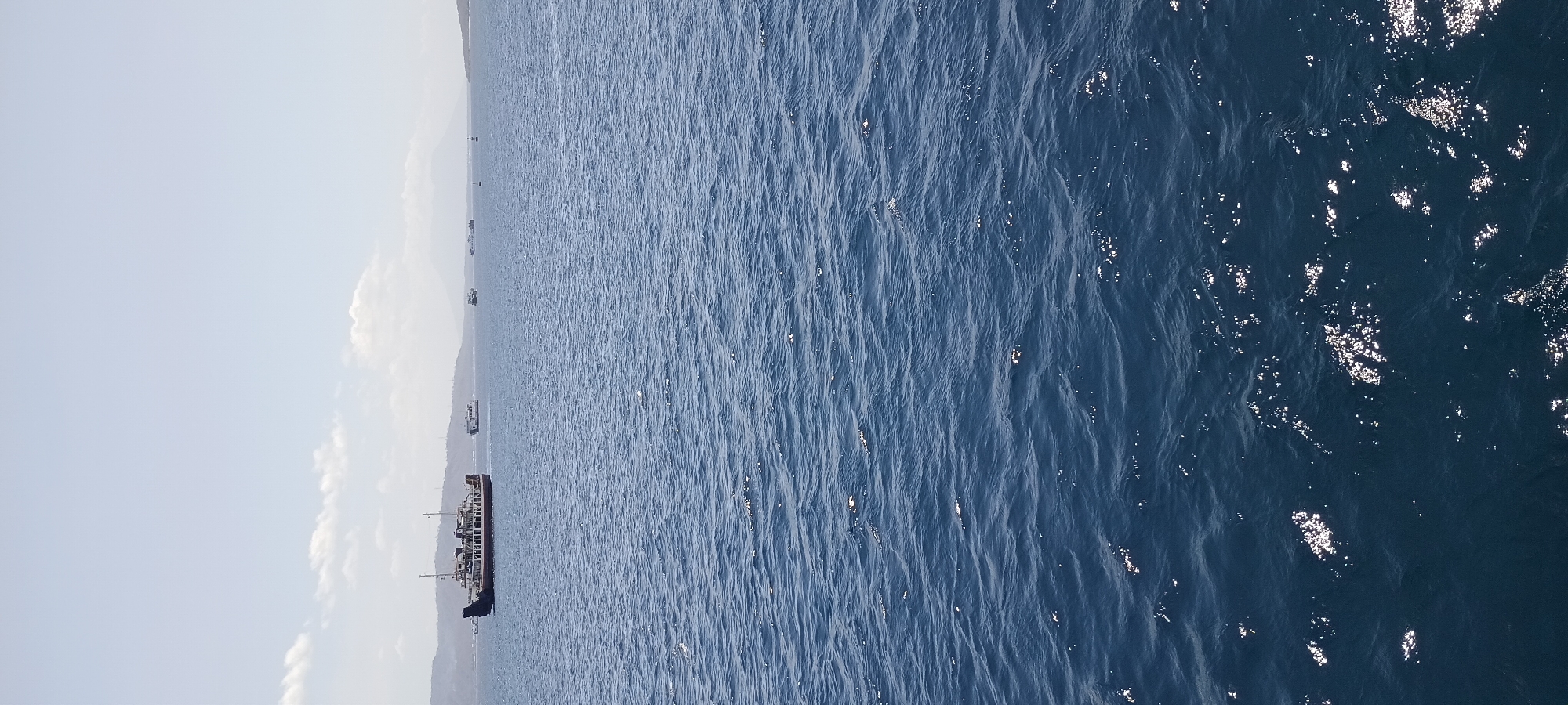 The image depicts a wide expanse of deep blue ocean under a pale sky with scattered clouds. Several ferries and boats are visible in the distance, navigating the calm waters. A large ferry, red and white, is the most prominent vessel, positioned on the left side of the frame. Further out, other ships and a distant landmass with hills and a mountain can be seen. The sun glints off the water's surface, creating bright highlights. The scene suggests a bright, clear day in a coastal area, likely Melaya, Indonesia, given the presence of ferries and the general geography.