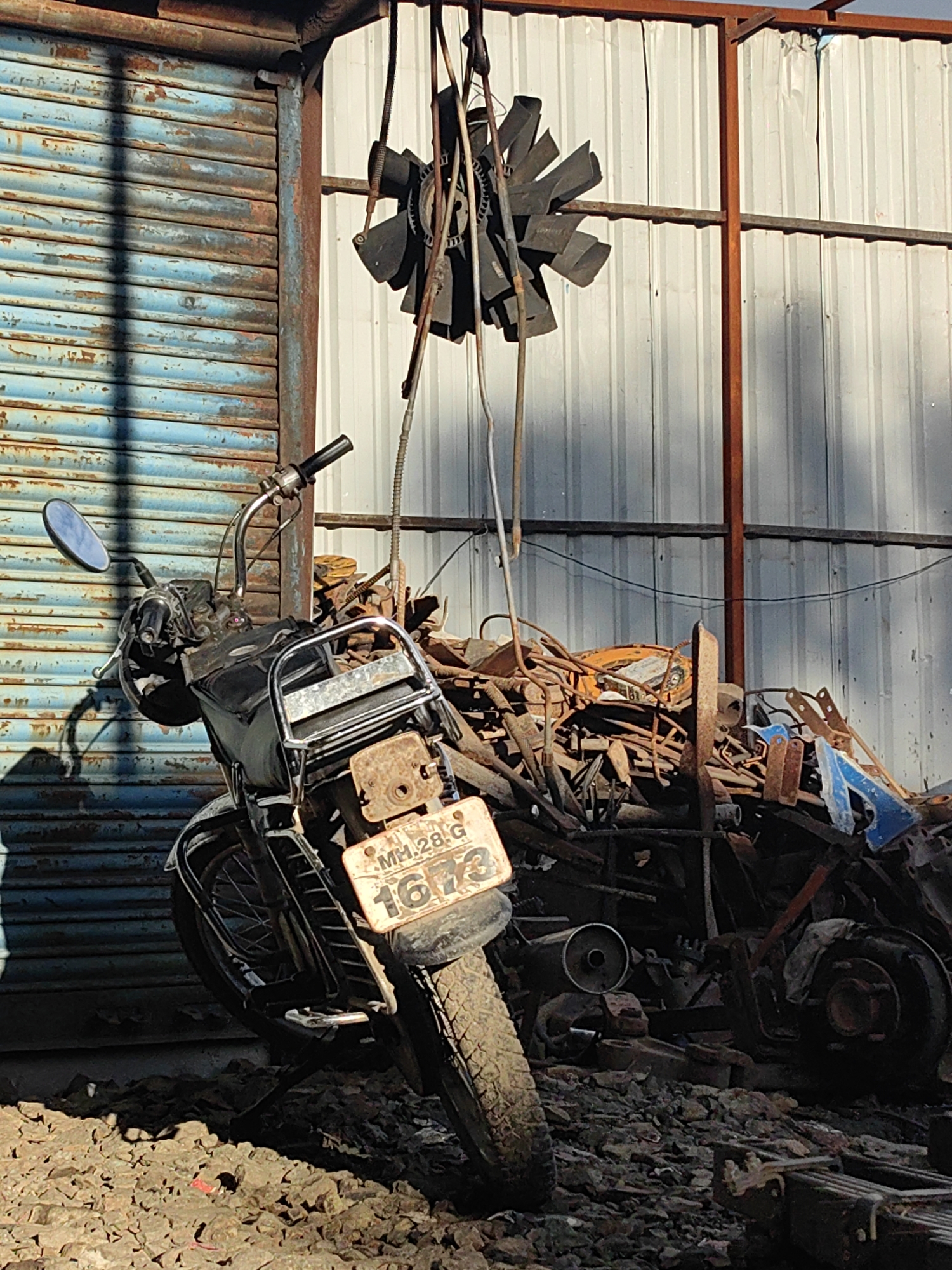 A motorcycle, its rear license plate reading "MH.28.G 1673", is parked in front of a rusty, blue corrugated metal roll-up door. Behind the motorcycle, a large pile of scrap metal is visible, composed of various rusted metal objects and some blue plastic. Hanging from a wire above the scrap pile is a black fan with several blades. The ground is covered in gravel and rubble. The scene appears to be an outdoor area in Malkapur, India, possibly a junkyard or workshop. There are no people visible in the image.