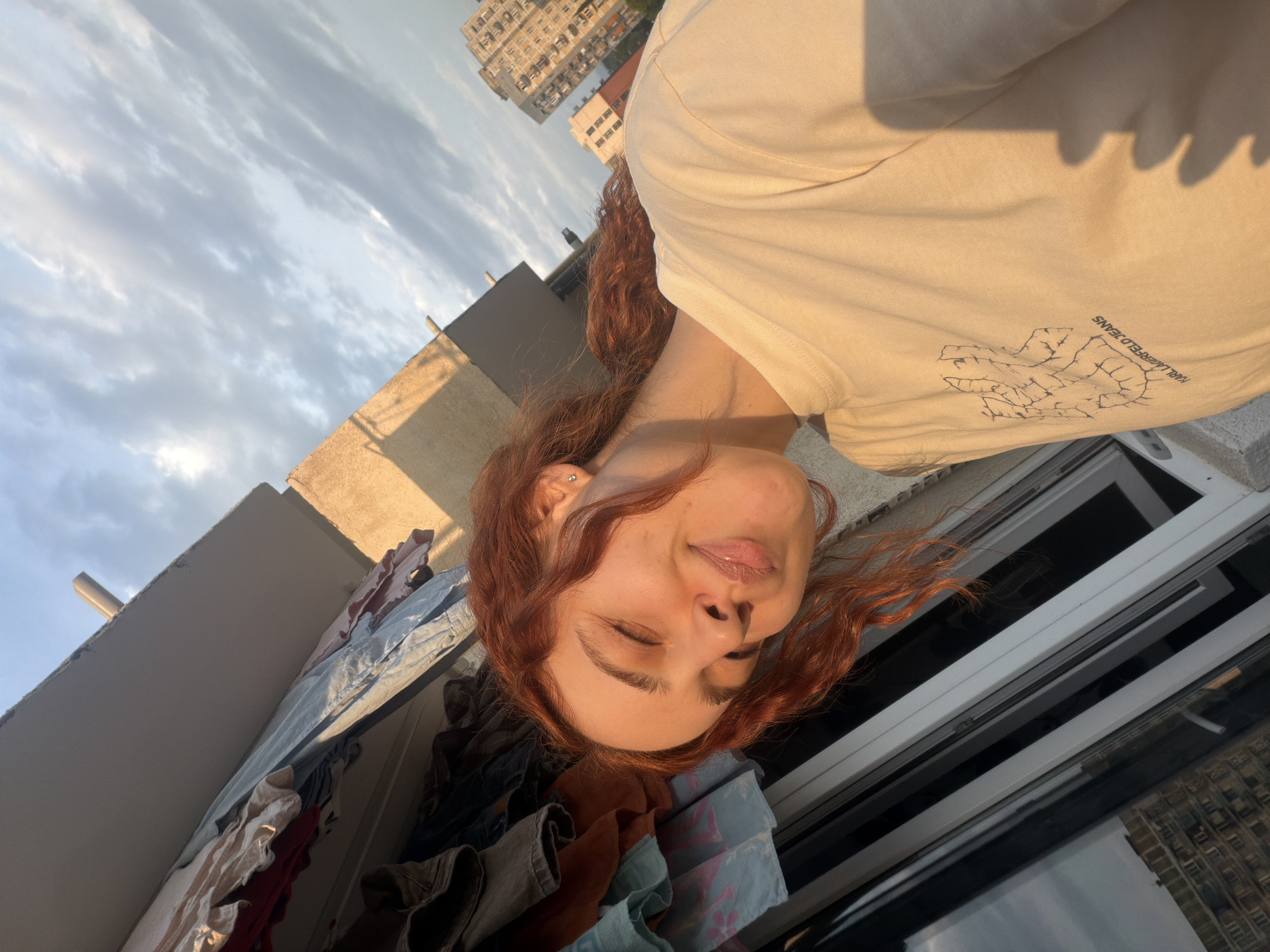 A red-haired person wearing a t-shirt, and located on an outdoor balcony or terrace in the city of Tbilisi, Georgia. Visible are buildings, cloudy skies, and laundry or clothes drying outside.