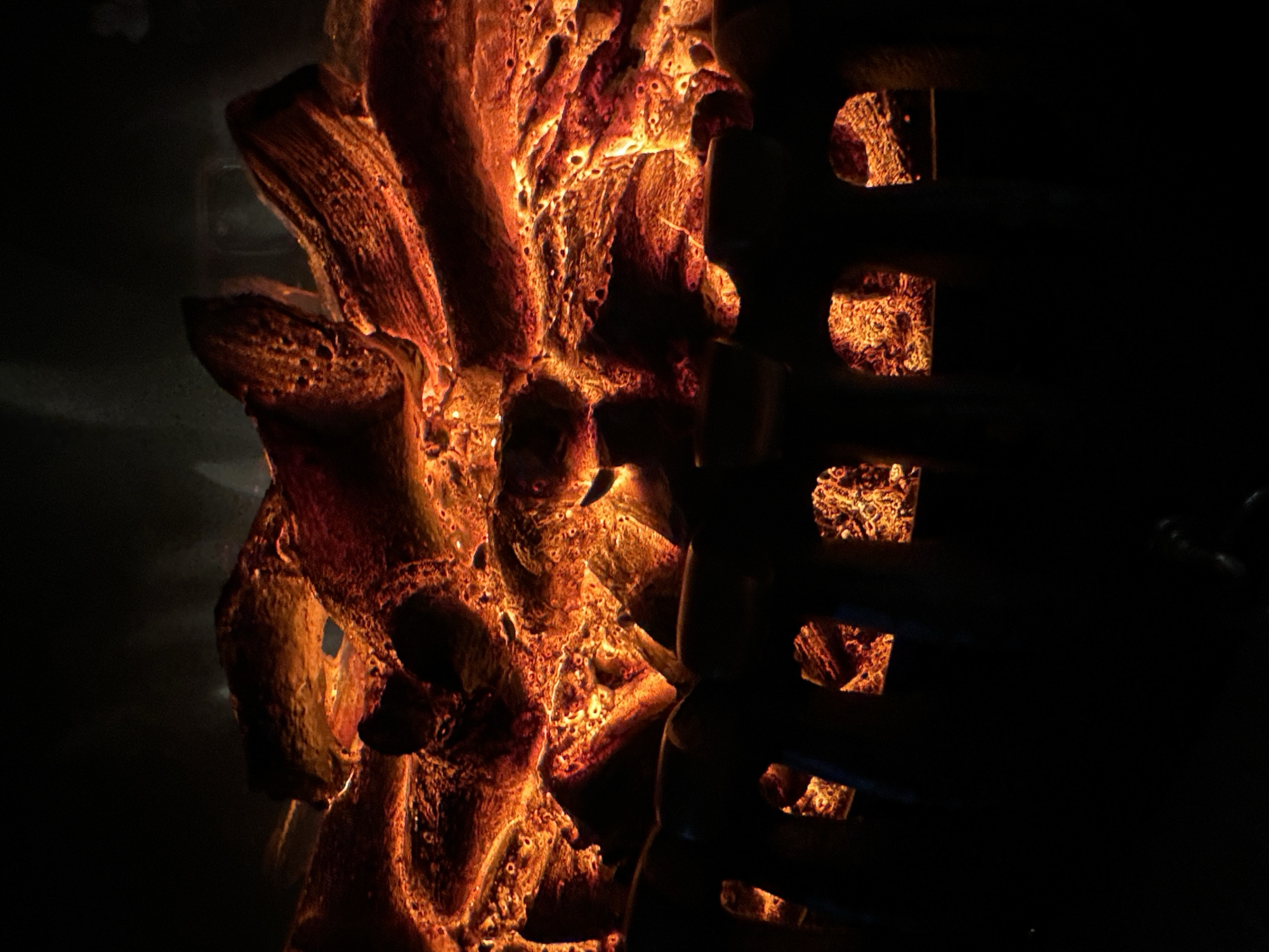 The image shows a close-up, low-angle view of an electric fireplace with glowing embers and artificial logs. The logs are textured and reddish-orange, with bright yellow and white highlights indicating heat and light. The embers are a vibrant mix of orange and red, creating a warm and inviting glow. Below the logs, a dark, shadowed metal grate with arched openings is visible, through which more glowing embers can be seen. The background is dark and indistinct, emphasizing the fiery display in the foreground. The overall impression is one of warmth and simulated fire. The location is Unknown, Unknown.