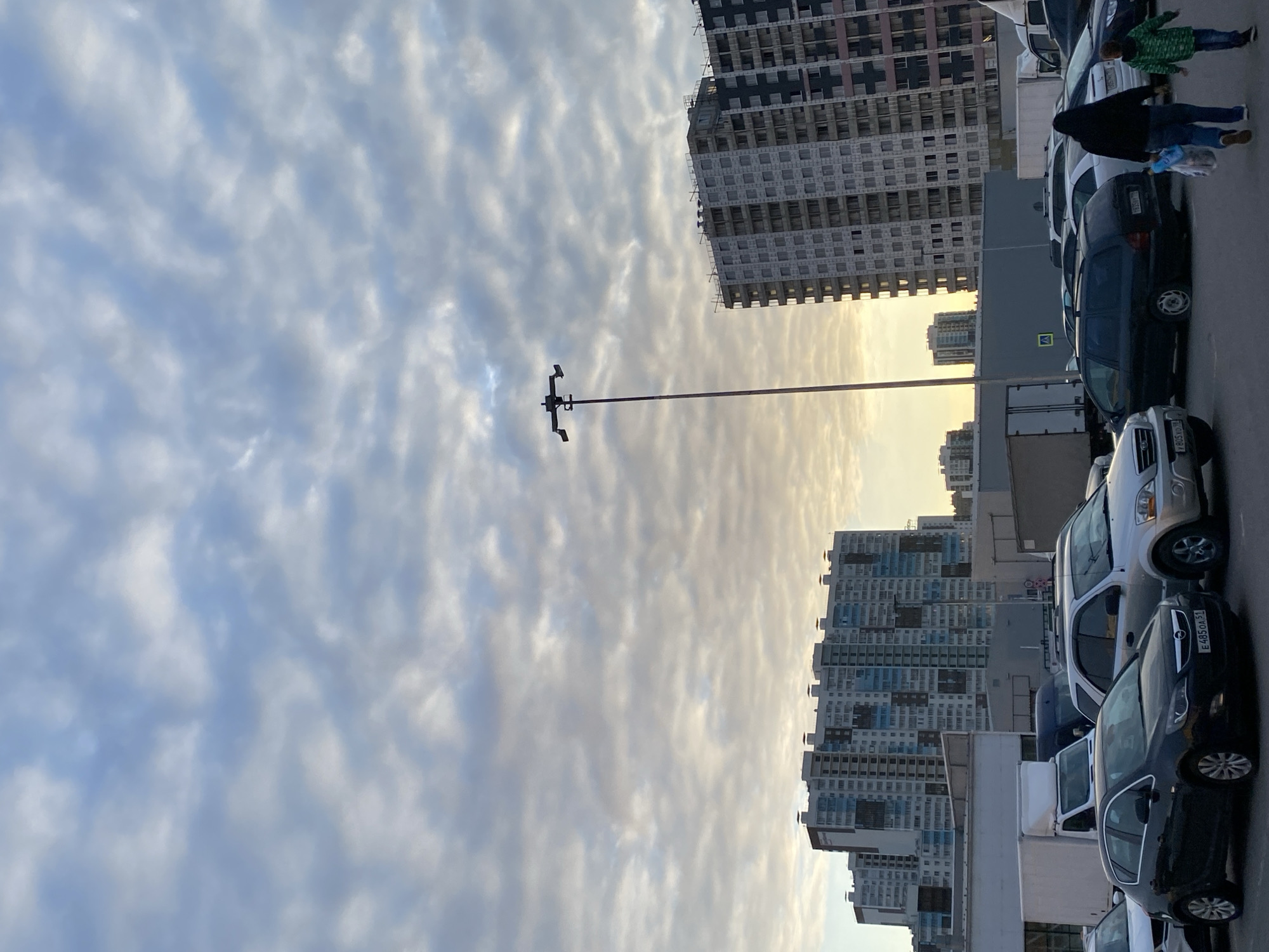 The image displays an urban landscape, captured from a rotated perspective, suggesting late afternoon or early evening due to the warm, soft light illuminating the horizon through a heavily clouded sky. The sky is a prominent feature, filled with numerous textured, white and grey clouds against patches of blue.
In the background, several tall, multi-story residential buildings stand, showcasing modern architecture with multiple windows and balconies. One building on the right appears to be under construction or recently completed, with visible scaffolding or netting on its exterior. Further in the distance, more buildings with varying facades contribute to the cityscape.
A row of parked vehicles, including sedans and SUVs, occupies the foreground on the right side. One dark sedan, possibly an Opel, bears a Russian license plate reading "E 485 OA 31." Another lighter SUV has a partially visible plate, "P 803 YO." Two individuals are seen walking near the parked cars; one, likely an adult, is dressed in a dark hooded jacket and blue pants, carrying a light-colored object. Accompanying them is a smaller person, presumably a child, wearing a green patterned jacket and blue pants.
A tall, slender pole, equipped with multiple fixtures, stands prominently in the mid-ground, silhouetted against the bright sky. Behind the vehicles, a grey structure features a yellow triangular sign with a black pedestrian symbol, indicating a crossing. The overall scene portrays a typical residential or mixed-use area within a modern city, consistent with the context of Saint Petersburg, Russia.
