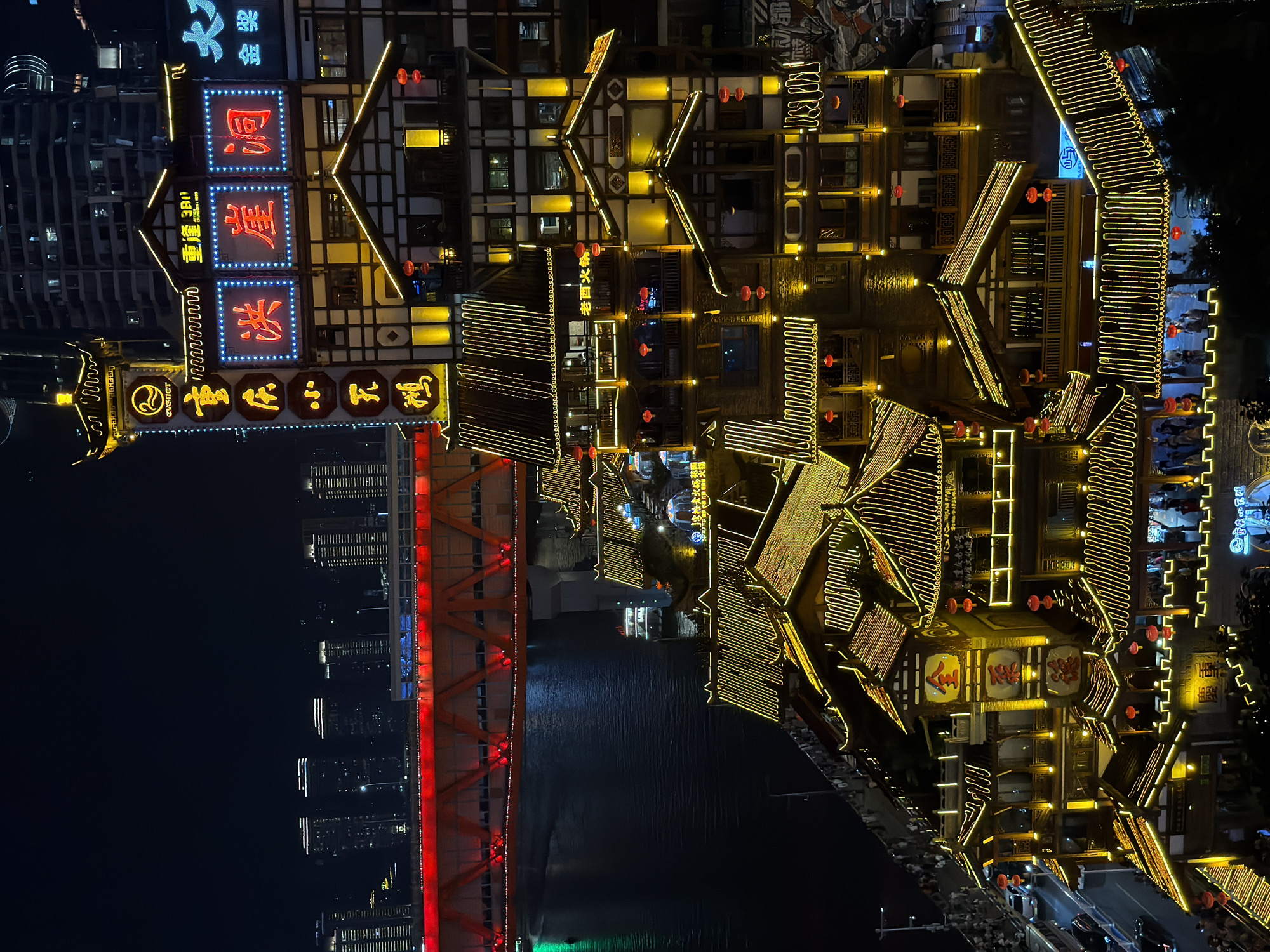 This is a nighttime aerial shot of the Hongyadong complex in Yuzhong District, Chongqing, China. The image showcases the multi-story, traditional Chinese architecture of the buildings, illuminated by a vast network of golden string lights outlining the roofs, balconies, and windows. Red lanterns are also visible, adding pops of color against the dark night sky. The Hongya Dong buildings are situated along the Jialing River, which is visible in the lower left of the frame, reflecting the lights. In the background, a bright red bridge spans the river, with modern skyscrapers lining the distant cityscape. The lower sections of the complex reveal glimpses of people walking along balconies and streets, with some cars visible on a road to the left. The scene is vibrant and bustling, with the traditional architecture standing in contrast to the modern urban environment.