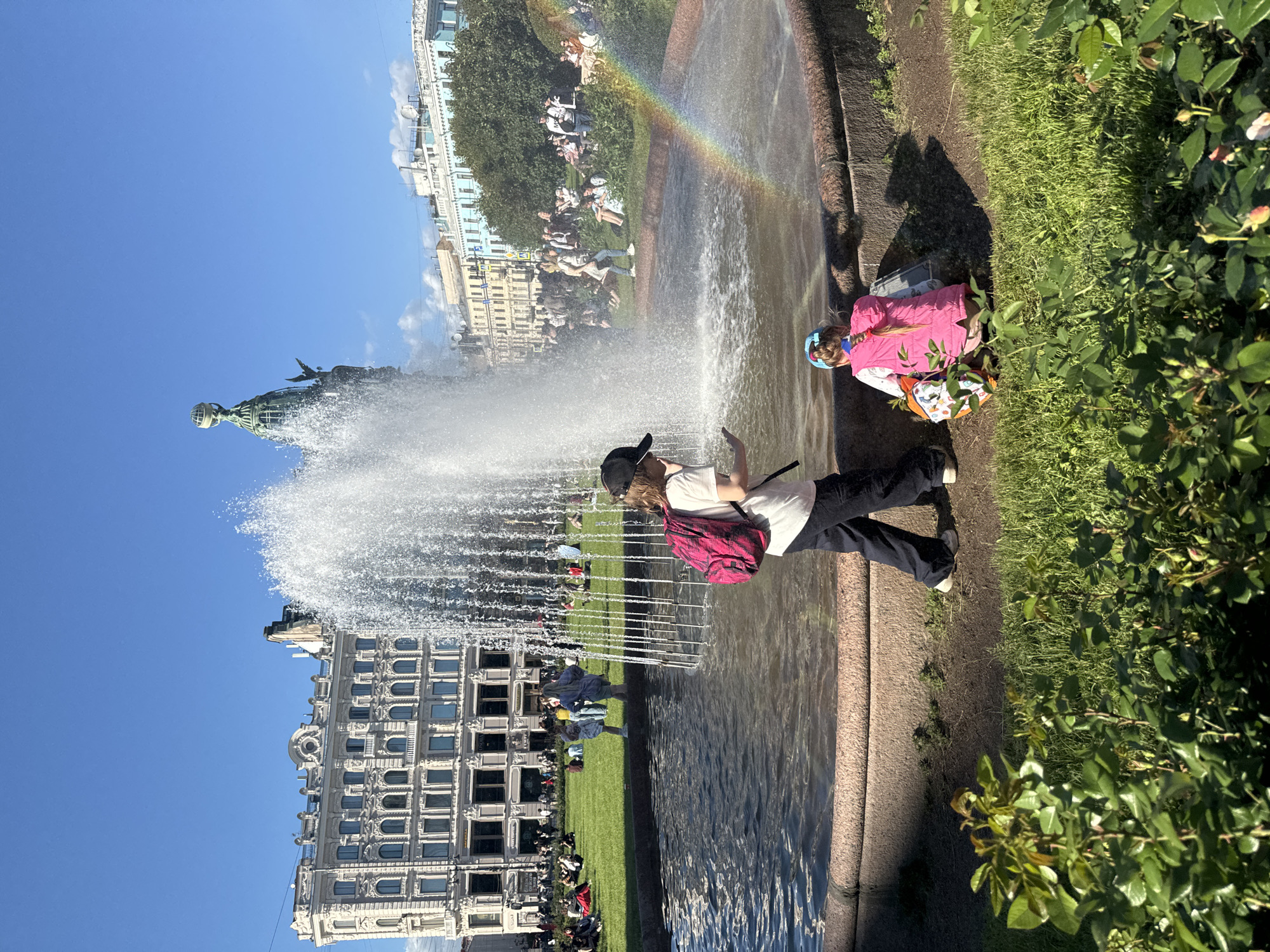 The image depicts a sunny day in Saint Petersburg, Russia, likely on a public square with a large fountain. In the foreground, two young children are near the fountain's edge. One child, facing away from the camera and wearing a pink vest and blue headband, is crouched down, seemingly playing or observing something on the ground. Another child, also facing away, is standing and appears to be holding a smartphone. The fountain is actively spraying water, and a subtle rainbow is visible in the mist.
In the mid-ground, a grassy area is dotted with people relaxing and sitting. Behind them, several large, ornate buildings with classical architecture line the square. One building in particular stands out with its detailed facade and arched windows. Further in the distance, trees and more buildings are visible under a clear blue sky. The overall atmosphere is one of a pleasant, leisurely day in an urban setting.