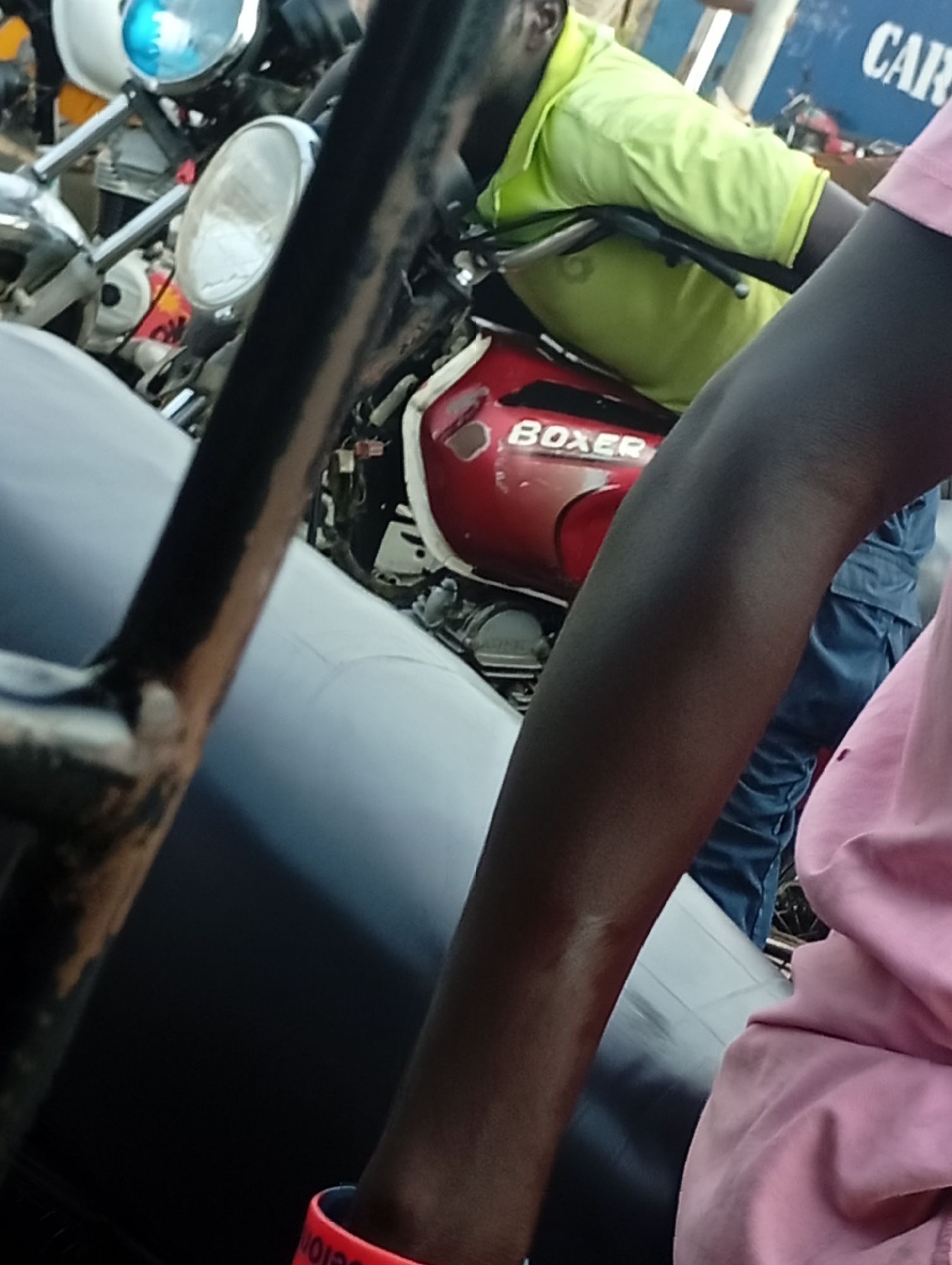 The image captures an outdoor scene featuring multiple motorcycles and people in what appears to be a street or open area during the daytime.
In the foreground, a dark, upholstered surface, possibly a vehicle seat, dominates the lower left. To its right, a dark-skinned arm in a pink long-sleeved shirt is visible, wearing a red wristband with partial text, possibly "ELON".
The midground features a person wearing a bright lime-green short-sleeved shirt, straddling or seated on a red motorcycle. This motorcycle clearly displays the word "BOXER" on its fuel tank. The person's hands are on the handlebars. Another person, partially visible in blue trousers, stands or sits near the red motorcycle. Behind and to the left of the "BOXER" motorcycle, parts of a white motorcycle are visible, including a headlight and a prominent blue light, possibly indicating an emergency or official vehicle.
In the background, a blue wall or large sign is discernible, with the partial word "CAR" written in white. The lighting suggests it is daytime. The overall setting appears busy, typical of a street scene or a place where vehicles are parked.