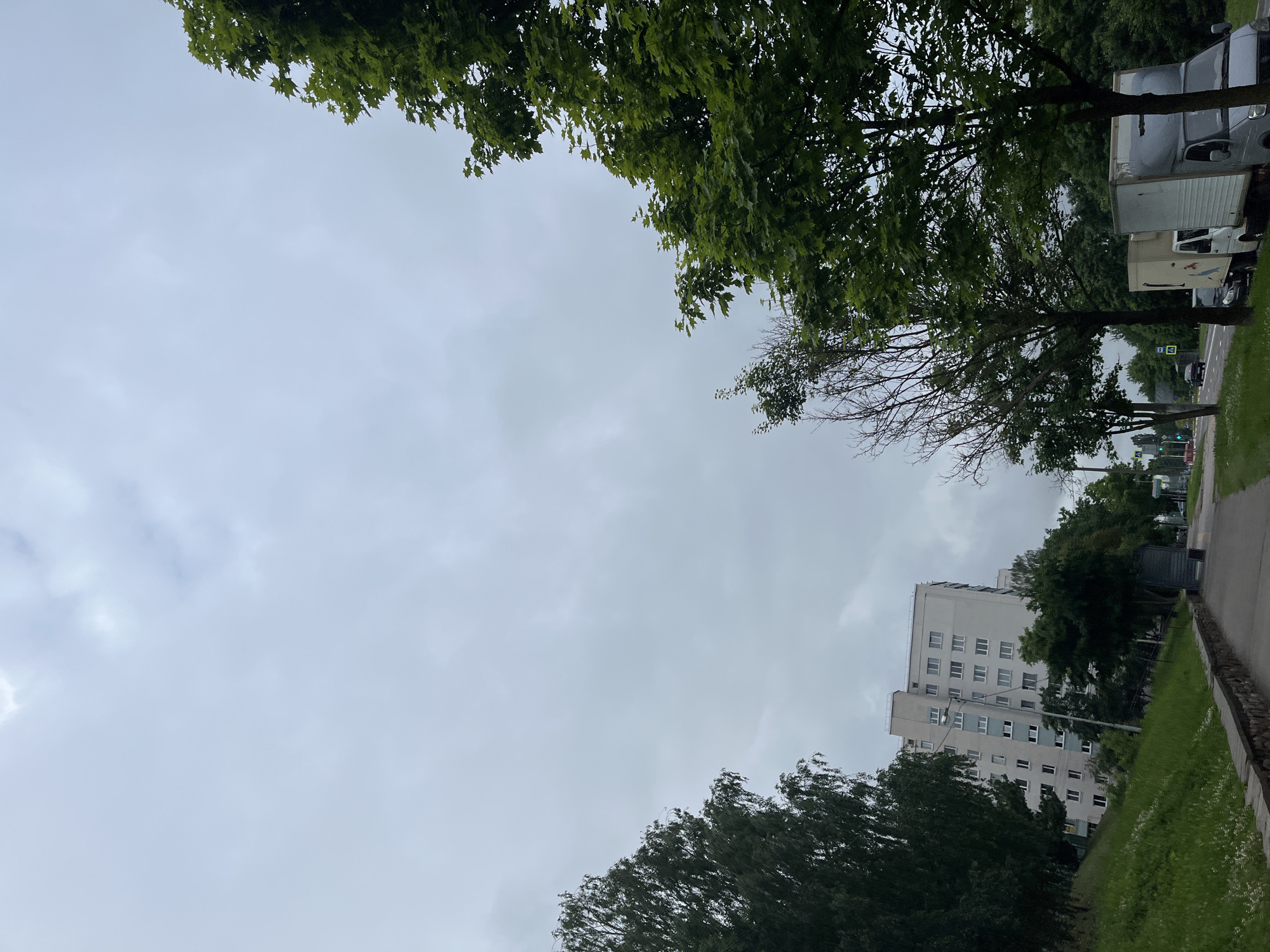 The image shows a cloudy sky over a street scene in Minsk, Belarus. On the left, a hill covered in green grass slopes down to a paved walkway. A multi-story, light-colored apartment building with numerous windows is visible on the slope. A dark-colored truck is parked on the street. On the right side of the walkway, several trees with green foliage are present, lining the street. Parked along the street are two trucks; one white box truck and a gray truck with a larger cargo box. Further down the street, traffic lights and other vehicles can be seen. The overall atmosphere suggests a typical urban environment on an overcast day.