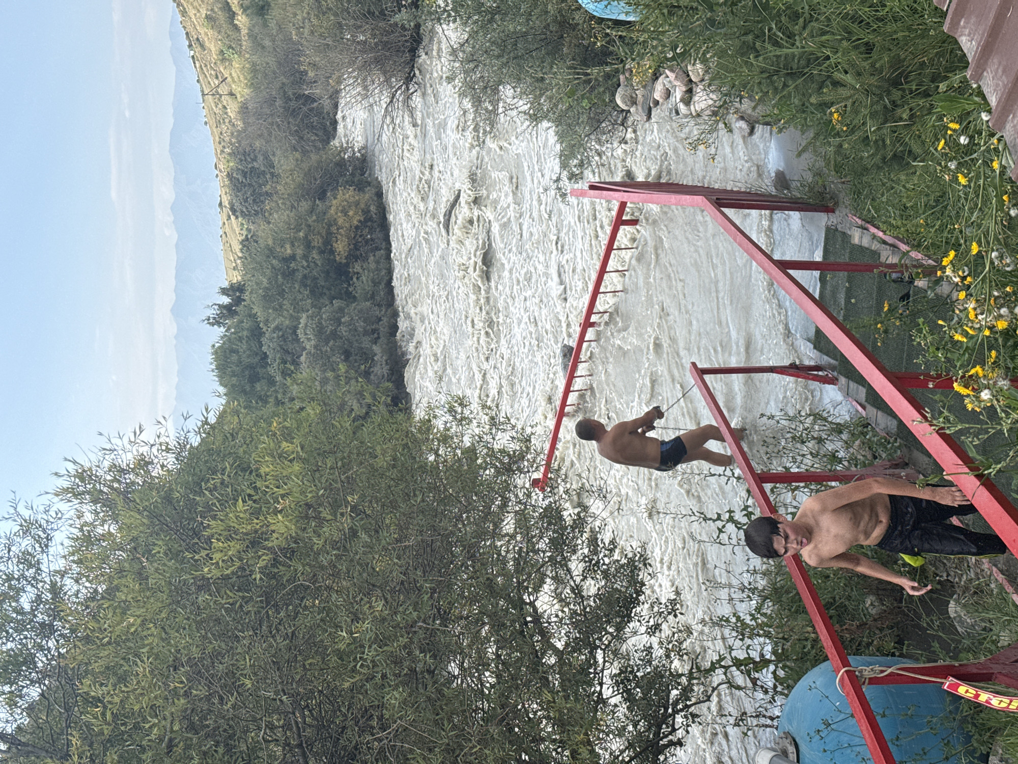 In Ak-Suu, Kyrgyzstan, a fast-flowing, muddy river dominates the scene. Two shirtless males are present. In the foreground, a young boy, appearing to be in his early teens, descends a set of stairs with red railings. He is wearing black shorts with yellow stripes and has a surprised or distressed expression. Further upstream, an adult male, wearing dark swim trunks, stands in the turbulent water, holding a long pole with a red rake-like attachment, possibly a tool for clearing debris or for a recreational activity. He is facing away from the camera. The riverbanks are lined with lush green trees and vegetation, with rolling hills and distant mountains under a light blue sky. The overall impression is a rugged, natural environment with human interaction.