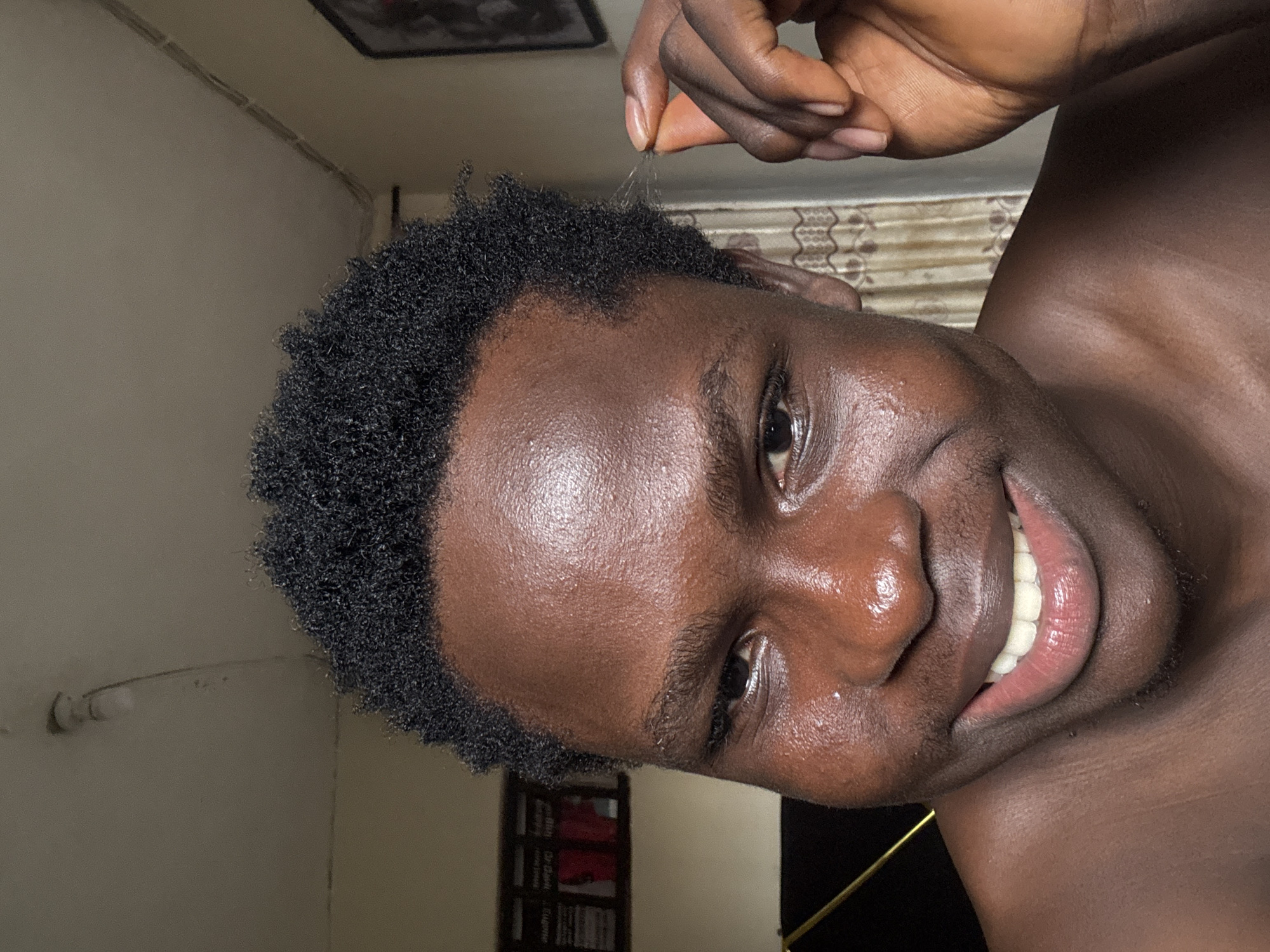 A close-up, head-and-shoulders shot focuses on the face of a young Black person with dark, curly hair and dark skin. Their eyes are open, and they are smiling, revealing white teeth. Their skin appears to be shiny, possibly from sweat or oil. The person is holding a few strands of hair between their thumb and index finger on the right side of the frame. In the background, a light fixture is visible on the ceiling, and a framed object hangs on the wall to the right, partially obscured by a patterned curtain. The lighting suggests an indoor setting.