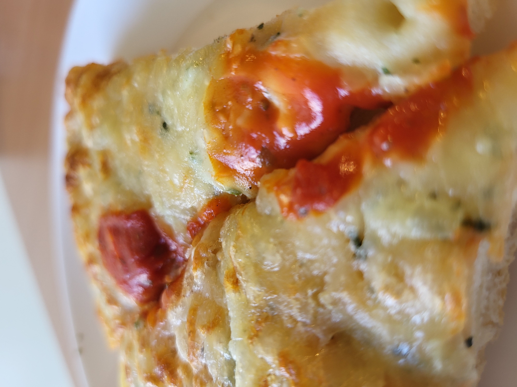 A close-up, angled shot shows a slice of pizza on a white plate. The pizza has a thick, golden-brown crust and is covered with melted white cheese. Dollops of red sauce are scattered across the cheese, some of which are partially melted and glistening. Small green flecks, likely herbs, are visible within the cheese. The background is blurred and appears to be a light-colored surface, possibly a table. The image is focused on the texture and details of the pizza.