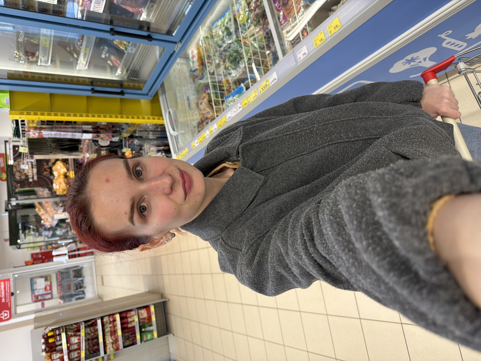 The image shows a woman taking a selfie inside what appears to be a supermarket in Chaikovsky, Russia. She has red hair and is wearing a gray sweater. She is holding a shopping cart with a red handle. Behind her, there are shelves and freezers filled with various products. The floor is tiled. Price tags are visible on the shelves.