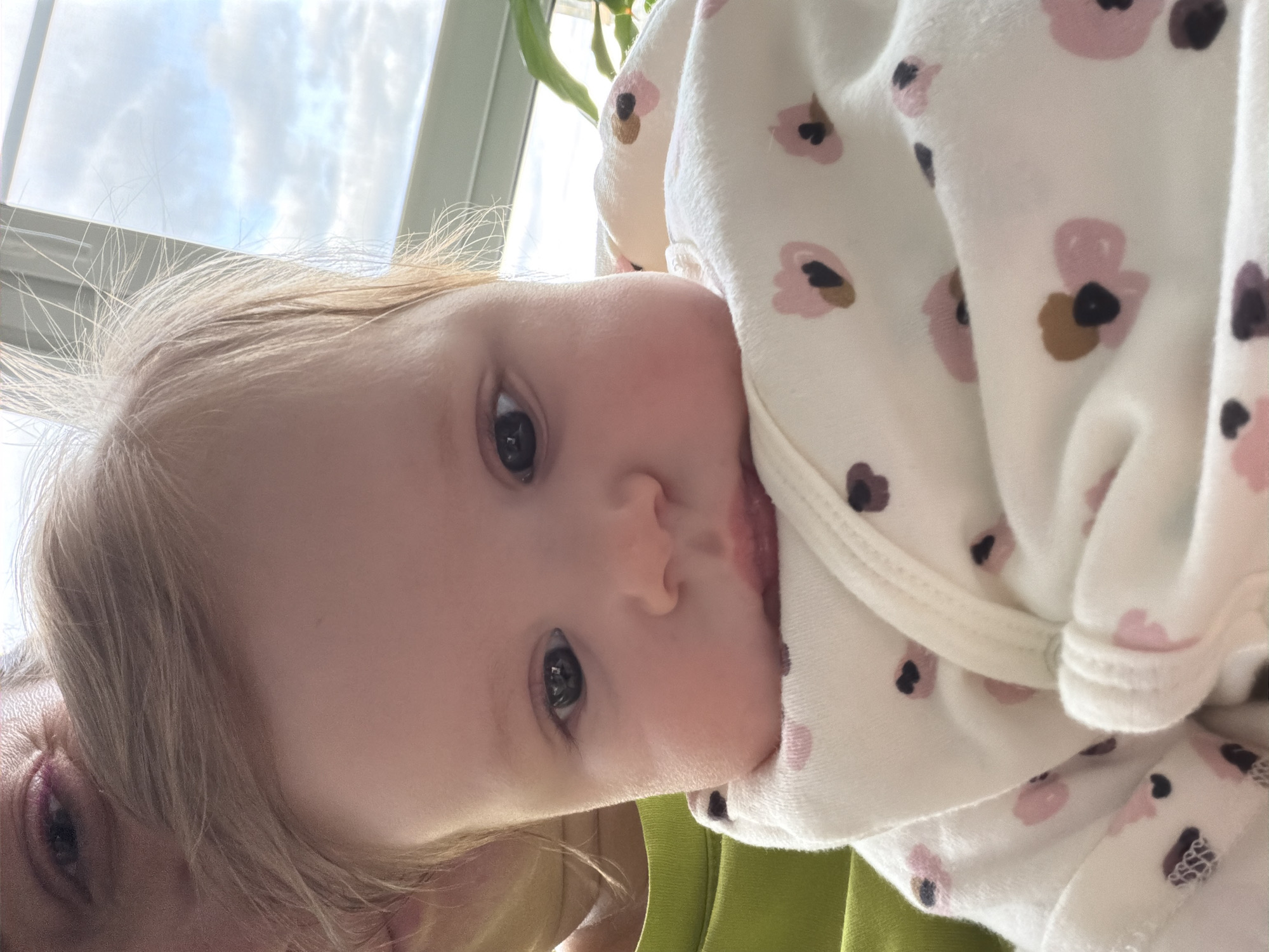 A close-up, slightly tilted photograph shows a baby and part of an adult's face. The baby has fair skin, light-colored hair, and dark eyes. The baby is wearing a white onesie with a pattern of stylized flowers in pink and brown. Part of the adult's face, showing an eye and blond hair, is in the lower left corner. The background includes a window with a view of the sky and a plant. The location is unknown, including the city of Podolsk, Russia.