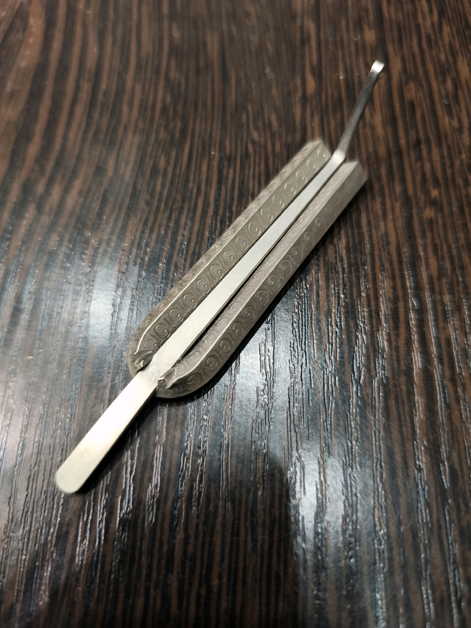 A close-up, overhead shot shows a metal jew's harp resting diagonally on a dark, wood-grained surface. The harp, a silver, oblong object with decorative etching along its sides, has a thin, flexible metal reed extending from its center. The surface beneath it is a rich brown with prominent, vertical wood grain lines, reflecting light in some areas, suggesting a polished or varnished finish. The lighting is soft, casting subtle shadows that define the object's contours. The scene is devoid of people or any indication of a specific urban environment.