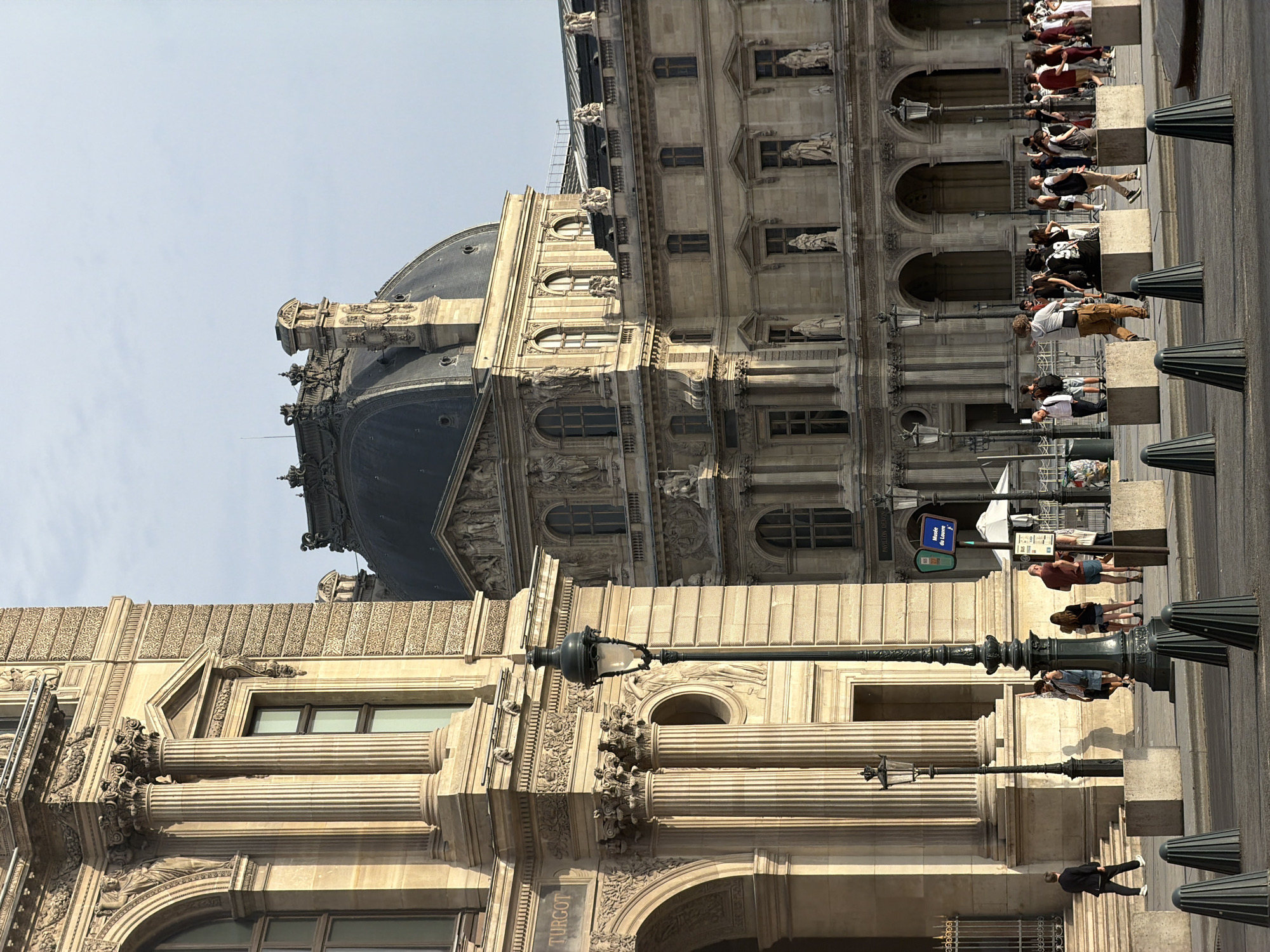 This is a photograph of the Louvre Museum in Paris, France, taken from the Cour Napoléon. In the foreground, a pedestrian street is visible, with concrete bollards lining the edge of the sidewalk. Several lampposts are scattered throughout the scene, casting shadows on the ground. A group of people are walking on the sidewalk, some of them appearing to be tourists. In the middle ground, the facade of the Louvre Museum is prominently displayed, featuring its iconic classical architecture with columns, arches, and ornate carvings. The large glass pyramid of the Louvre is not visible in this particular shot. In the background, the Parisian sky is a pale blue with some clouds. The lighting suggests it is either early morning or late afternoon, with the sun casting long shadows.