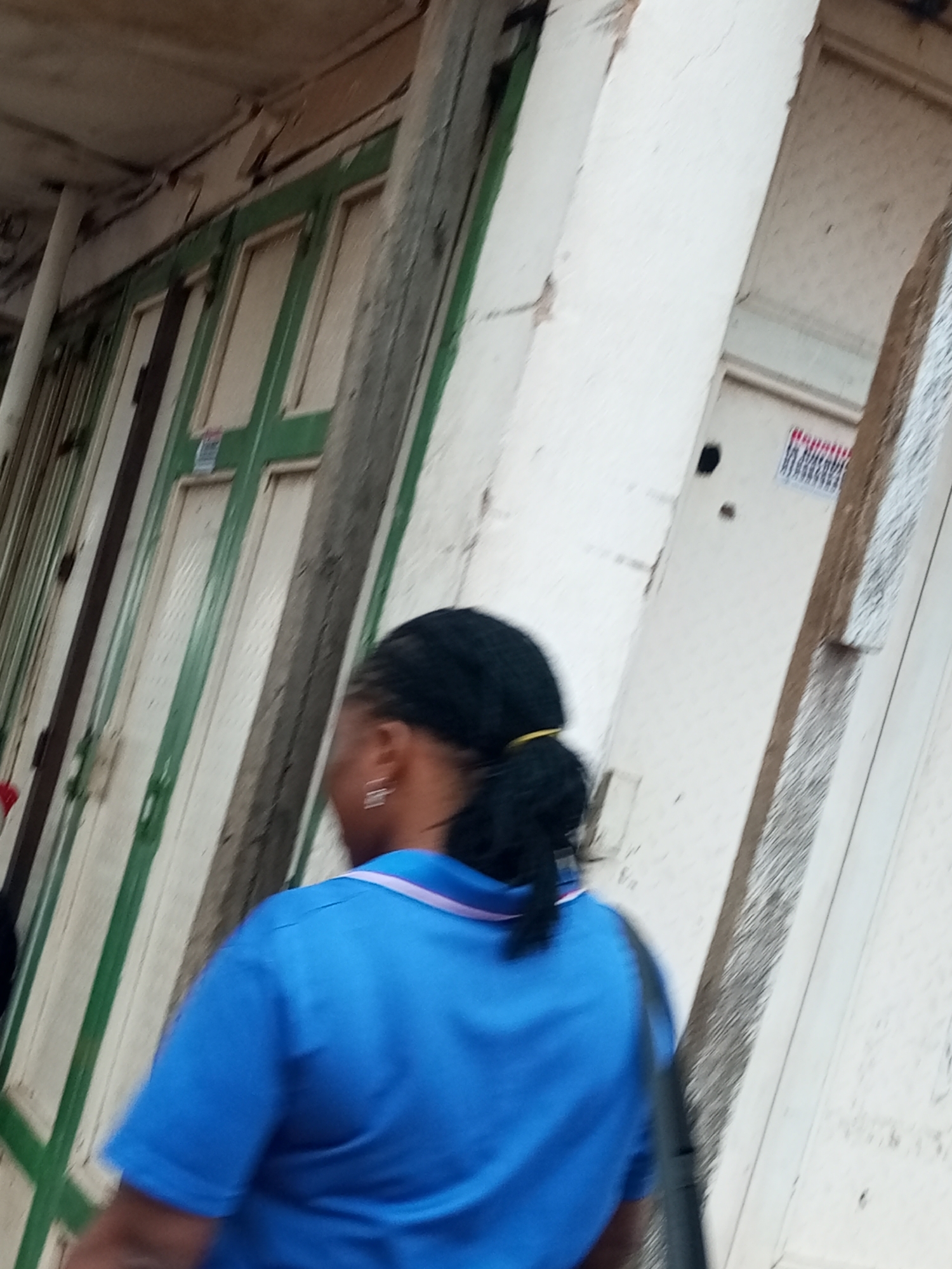 The image shows the back of a person wearing a blue t-shirt with a pink and white collar, and a black ponytail. The person is walking past several closed storefronts with green and white painted doors. A red and white sign is visible on one of the doors. The scene appears to be outdoors in a city location, possibly Tempure, Nigeria, judging by the architecture and style of the storefronts. The overall lighting suggests it is daytime.
